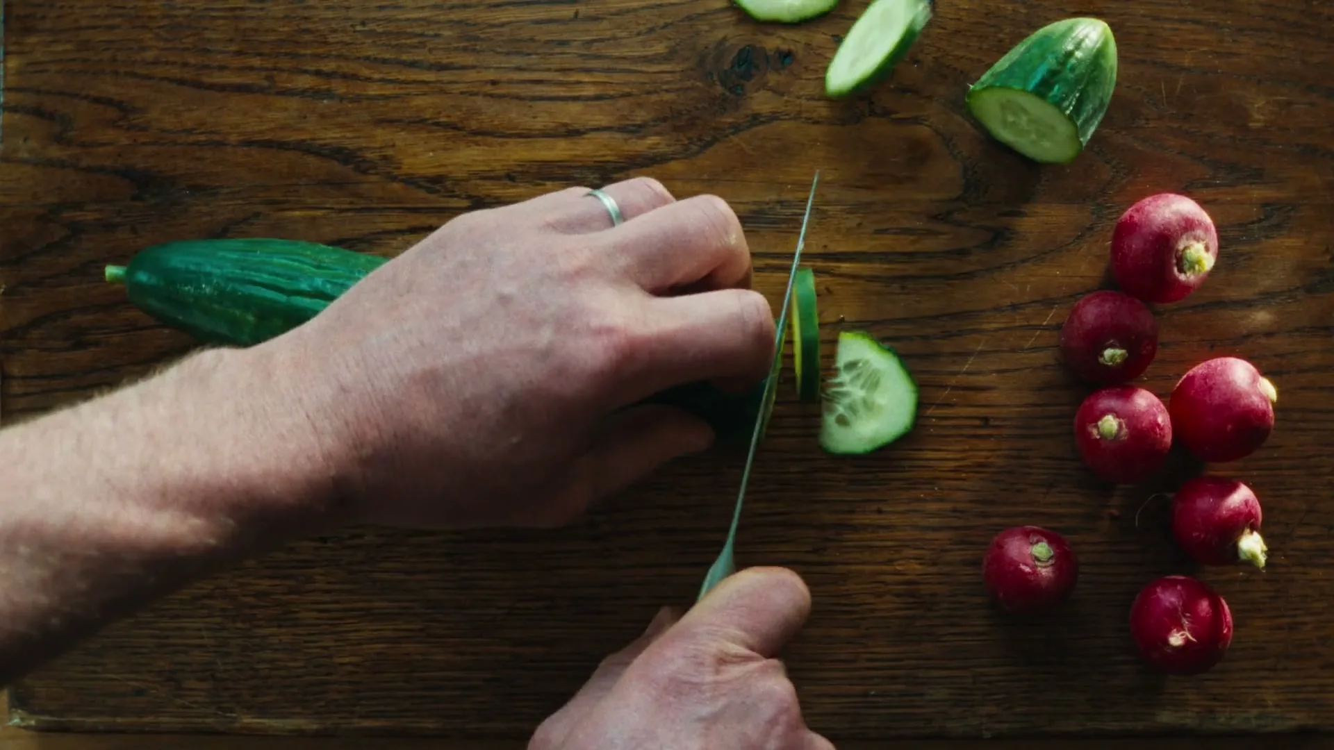 A person's hands are seen from an overhead perspective, slicing a cucumber on a wooden cutting board next to several red radishes.