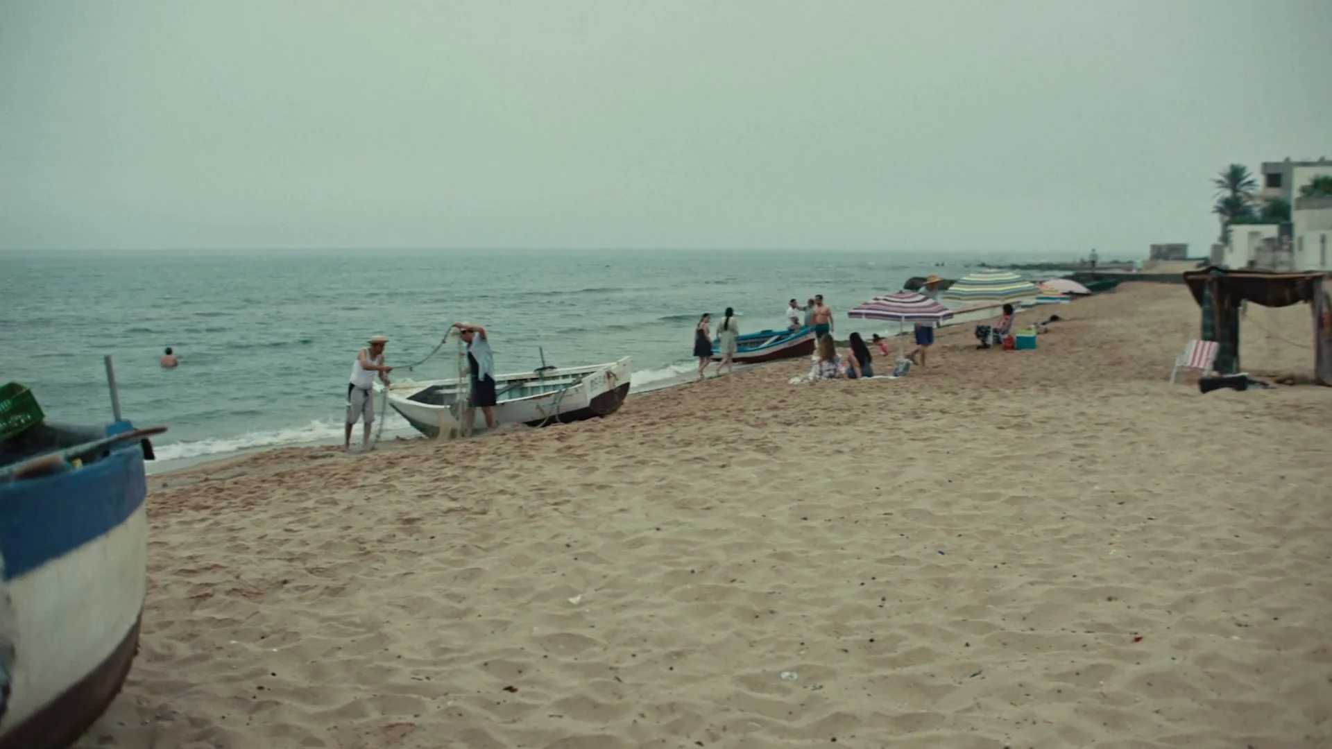 A group of children play soccer on a sandy beach, while adults relax and two fishermen work with their boats near the water.