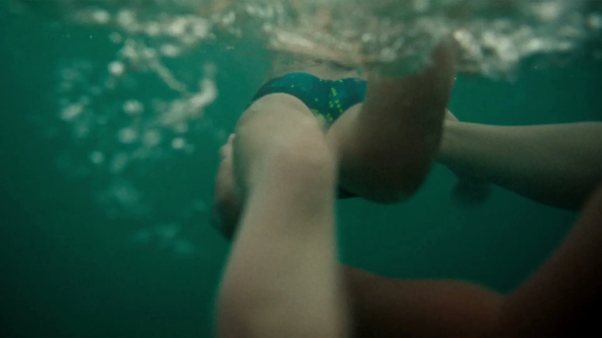 Underwater, a child in blue swim trunks is held by an adult's arm, then they surface, creating splashes.
