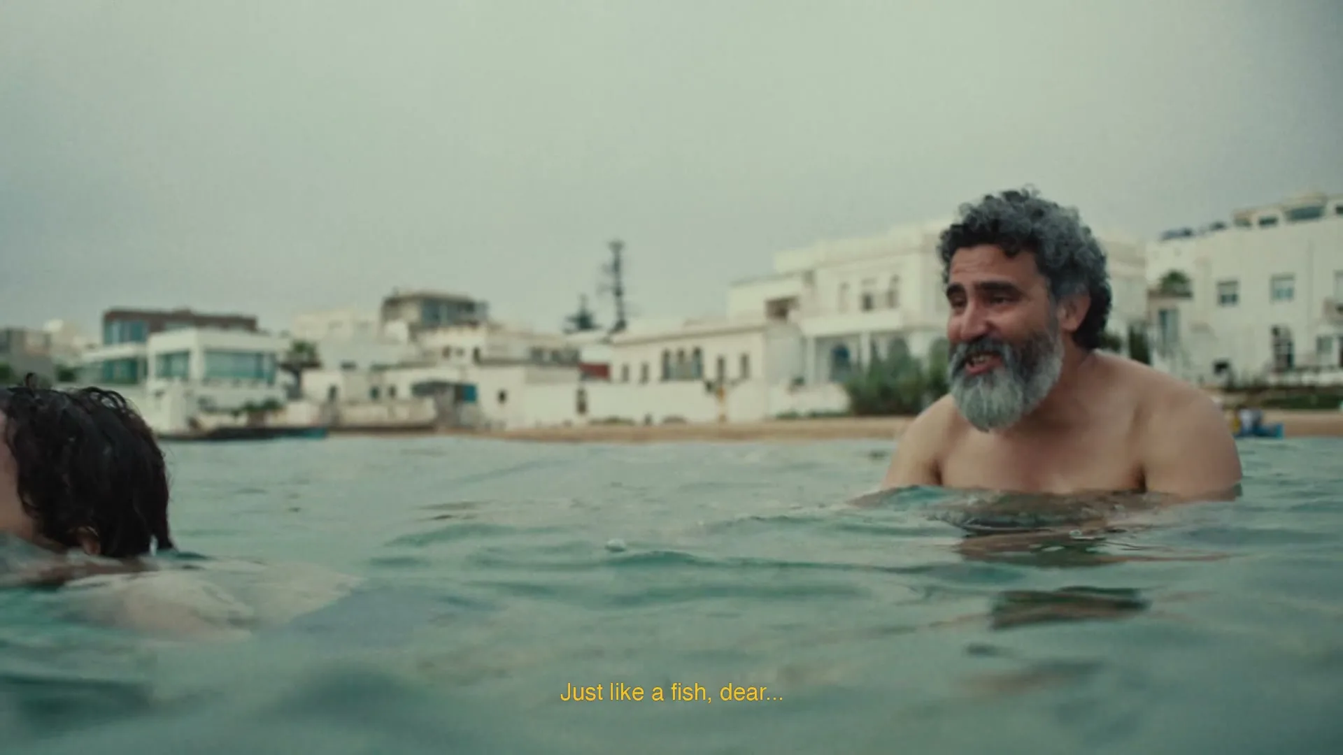 A man with a grey beard and a young girl swim in the ocean, with white buildings visible on the shore in the background.