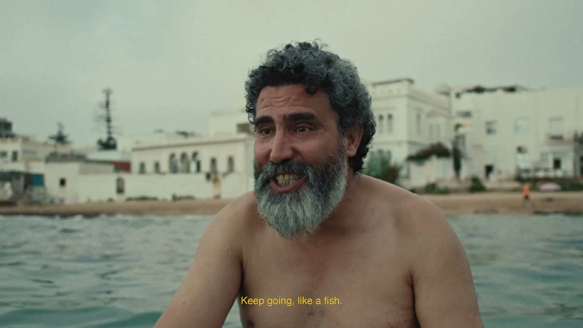 A middle-aged man with a grey beard and curly hair is in the water, looking at someone off-screen and talking, with buildings in the background.