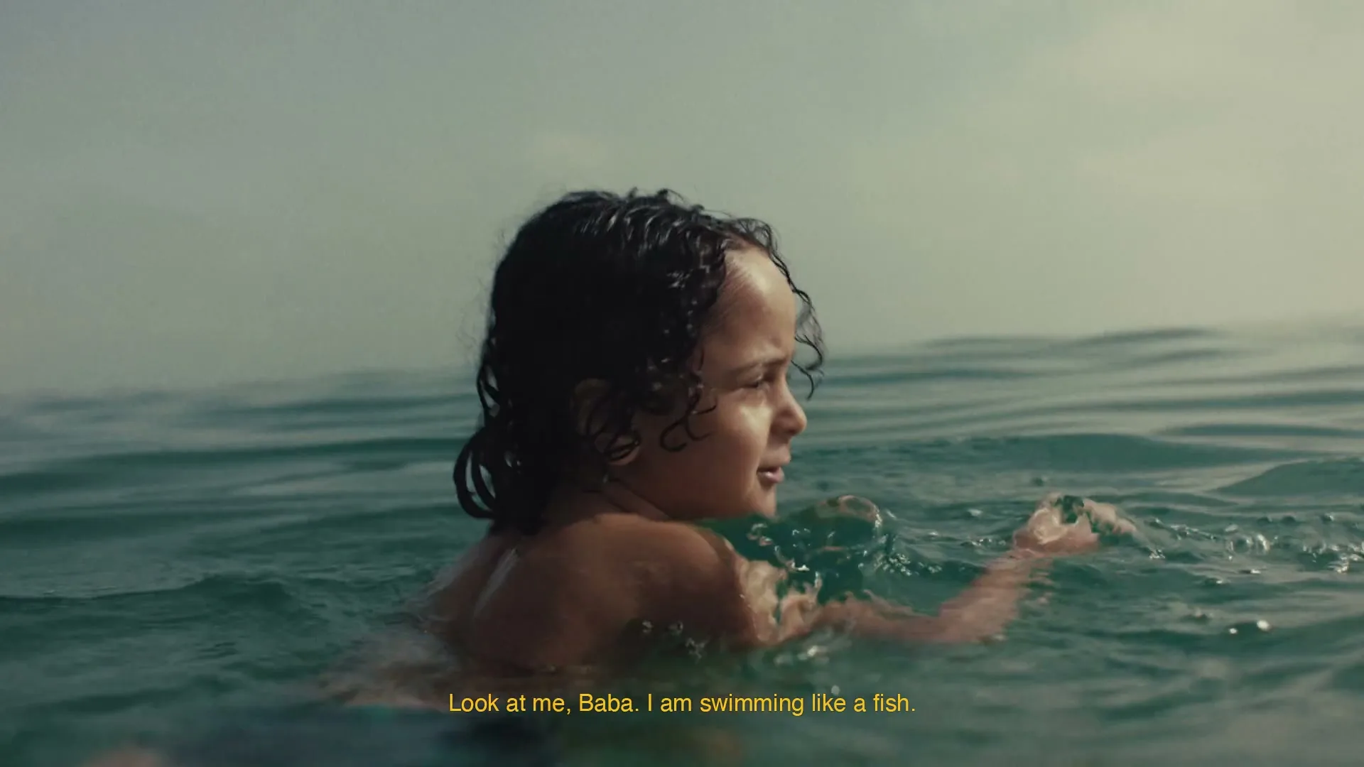 A young child with curly hair swims in the ocean, looking back over their shoulder with a hopeful expression as water splashes around them.