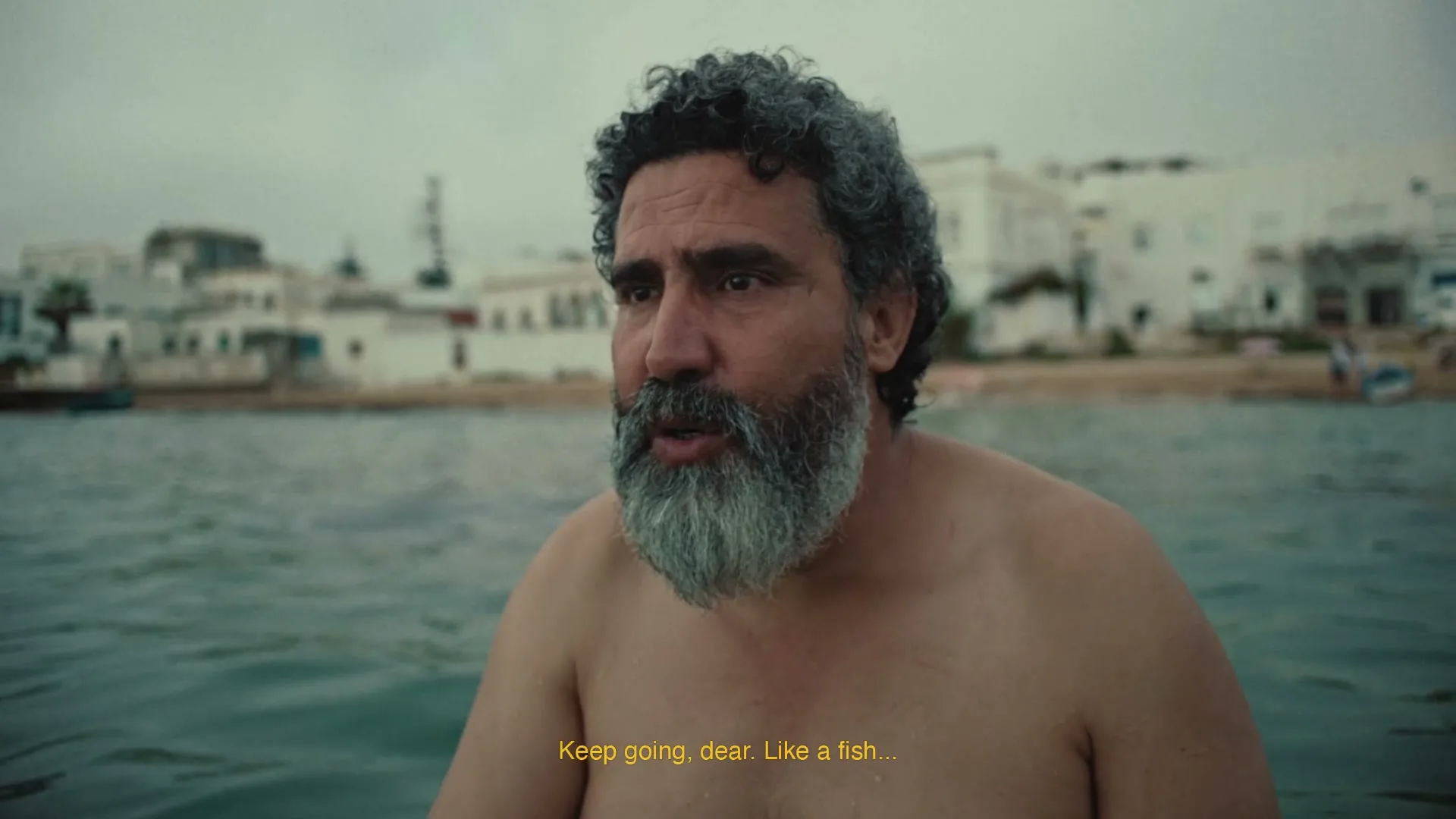 A middle-aged man with a grey beard and curly hair is in the water, looking towards the right side of the frame, with a coastal town in the background.