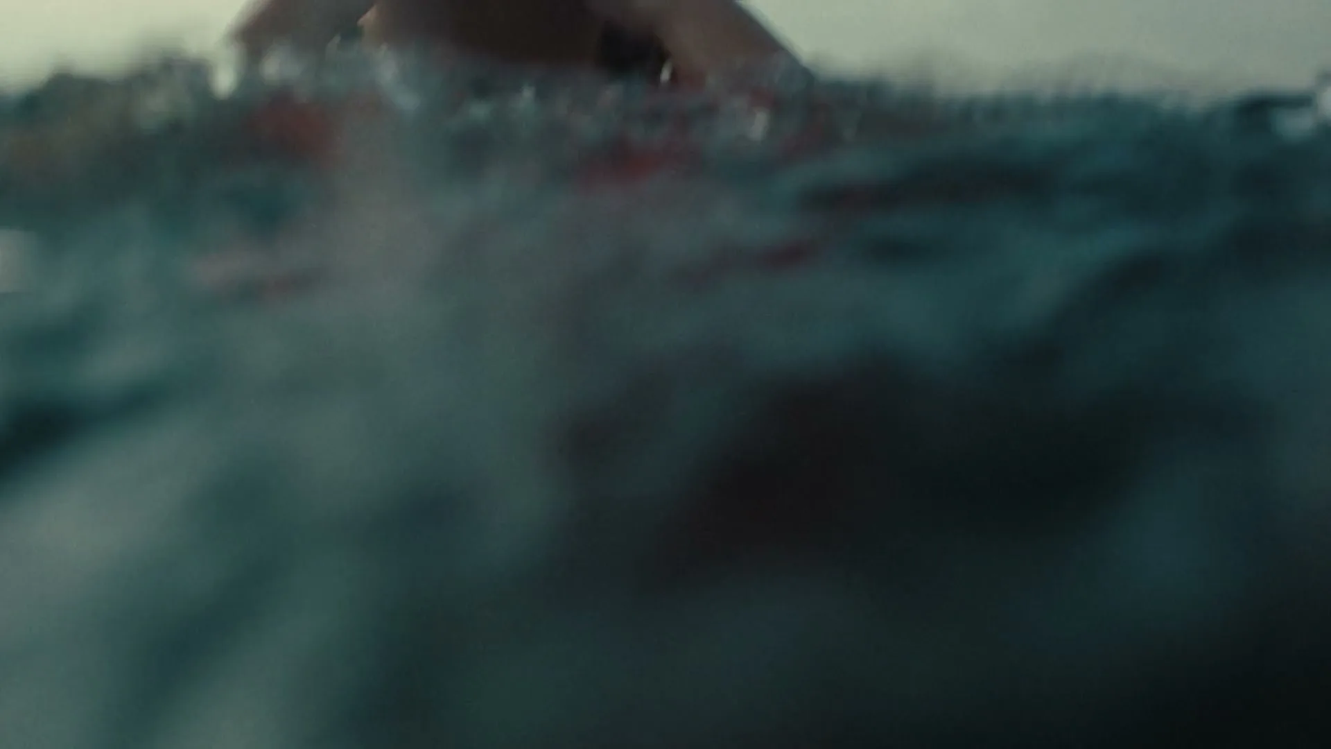 A young woman with wet hair and a red life vest emerges from the water, wiping her eyes, in the open sea.