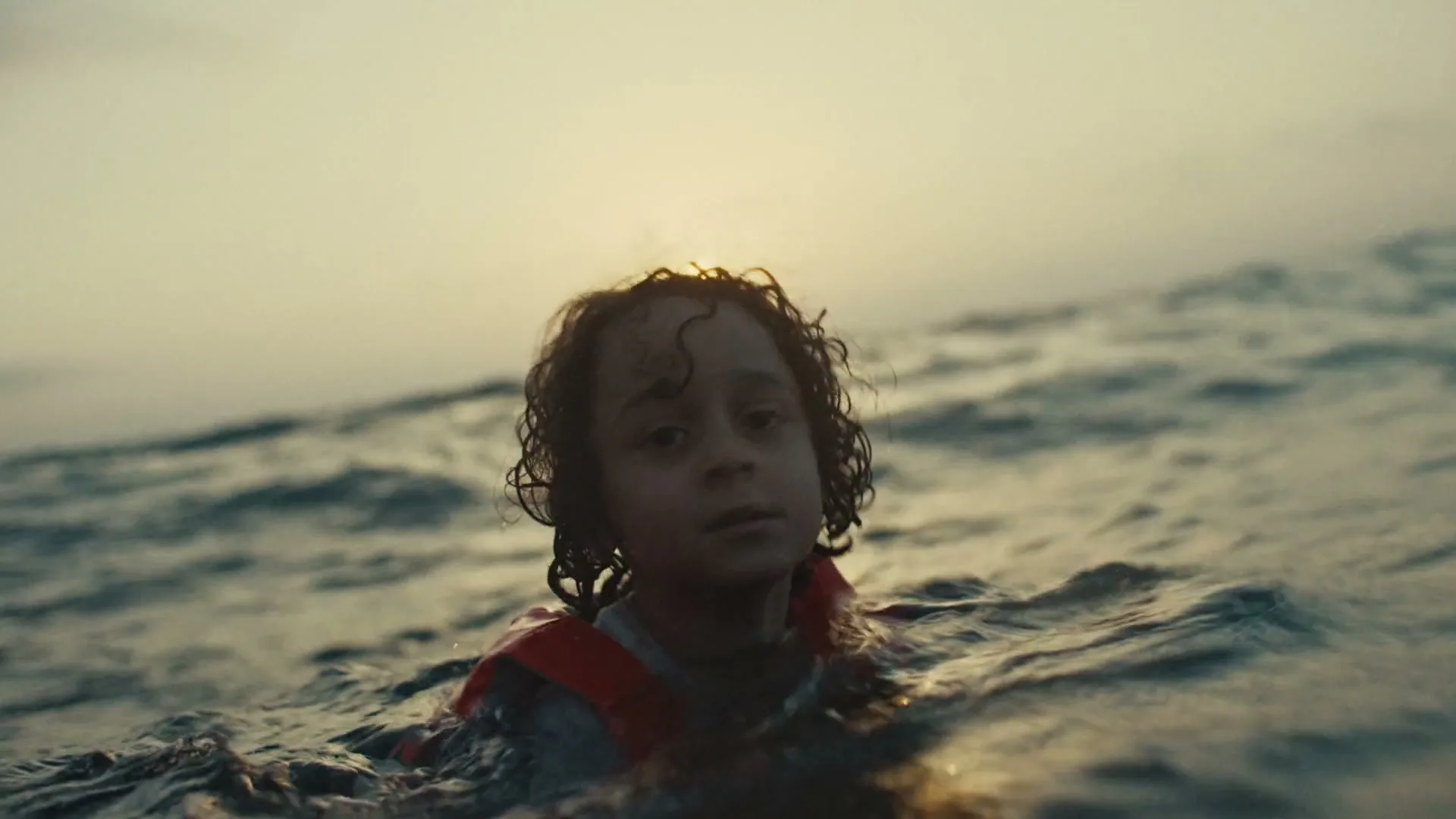 A young child with curly hair and a life vest floats in the ocean at sunset, looking around with a serious expression.
