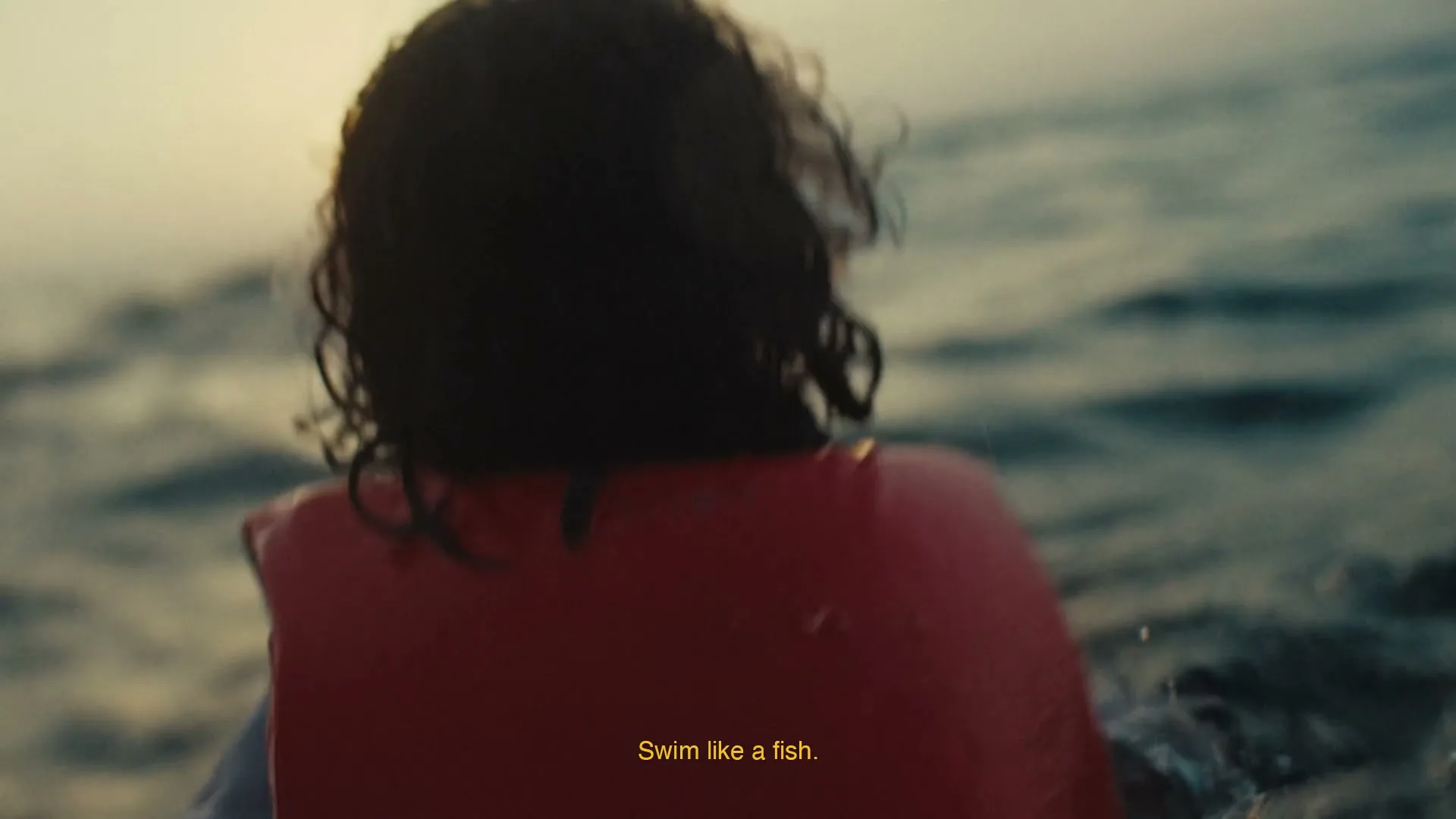 A person with dark, curly hair and a red life vest is seen from behind, struggling to swim in dark, choppy water at dusk.