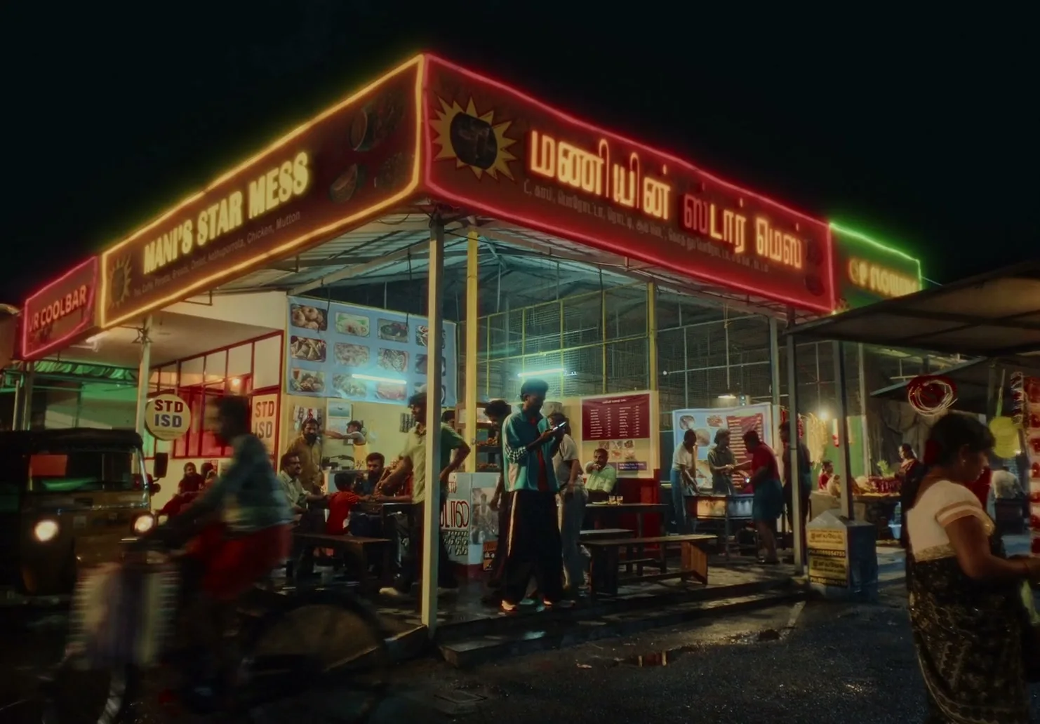 A wide shot of a bustling street food stall at night, with people eating and walking around under neon signs.