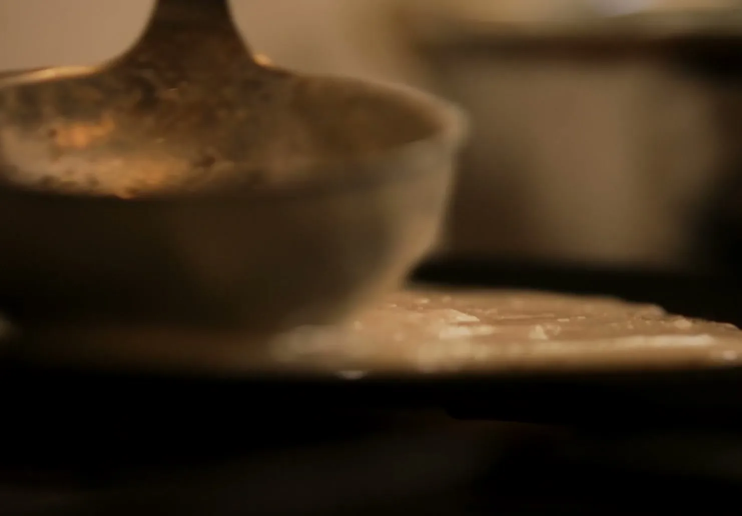 A close-up shot shows a ladle pouring liquid onto a flat surface, possibly cooking, in a dimly lit interior.