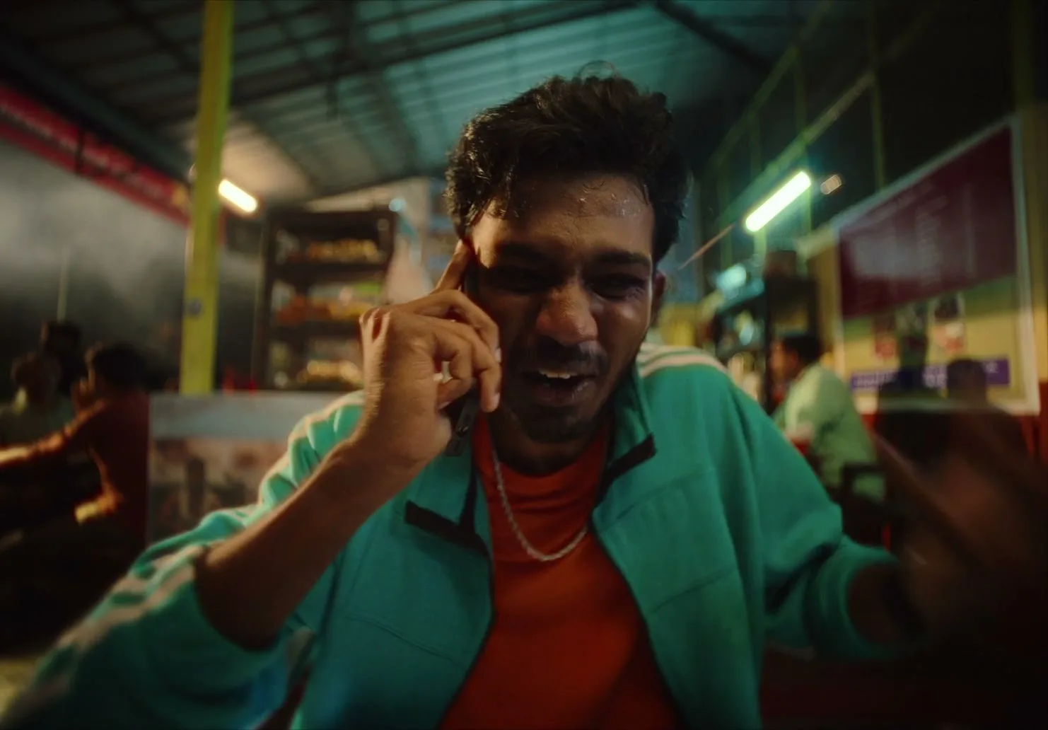 A young man in a teal jacket talks animatedly on a phone, then turns to face an older man in a yellow shirt who leans in close, in a bustling, dimly lit eatery.
