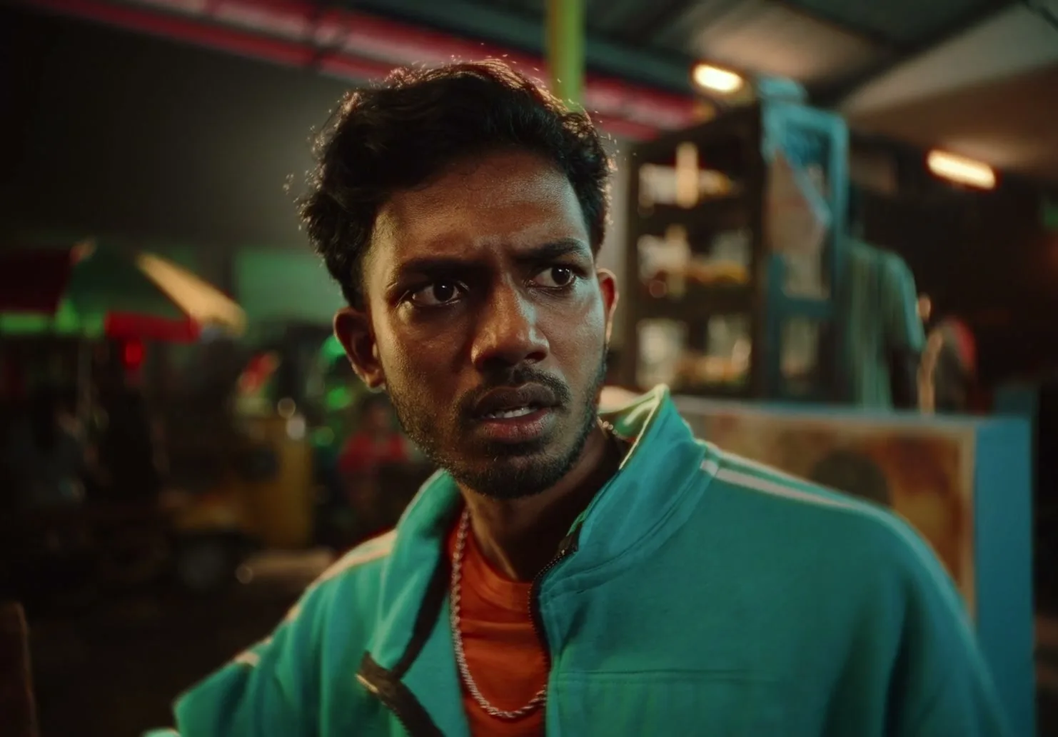 A young man with dark skin and a beard, wearing a teal jacket and orange shirt, looks around with a worried expression in a busy night market.