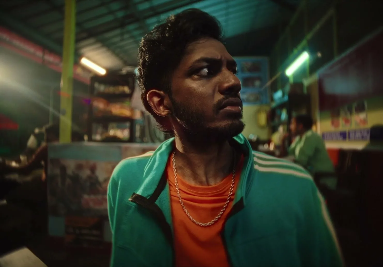 A young man with dark hair and a beard, wearing a teal jacket and orange shirt, looks around nervously in a busy, dimly lit restaurant.