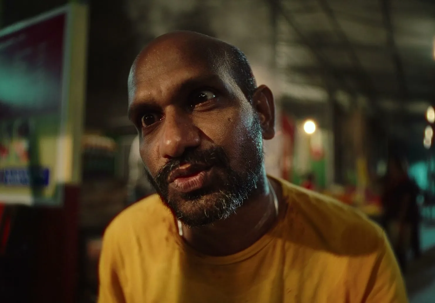 A bald, dark-skinned man with a beard and mustache, wearing a yellow t-shirt, looks up and speaks at night in a market.
