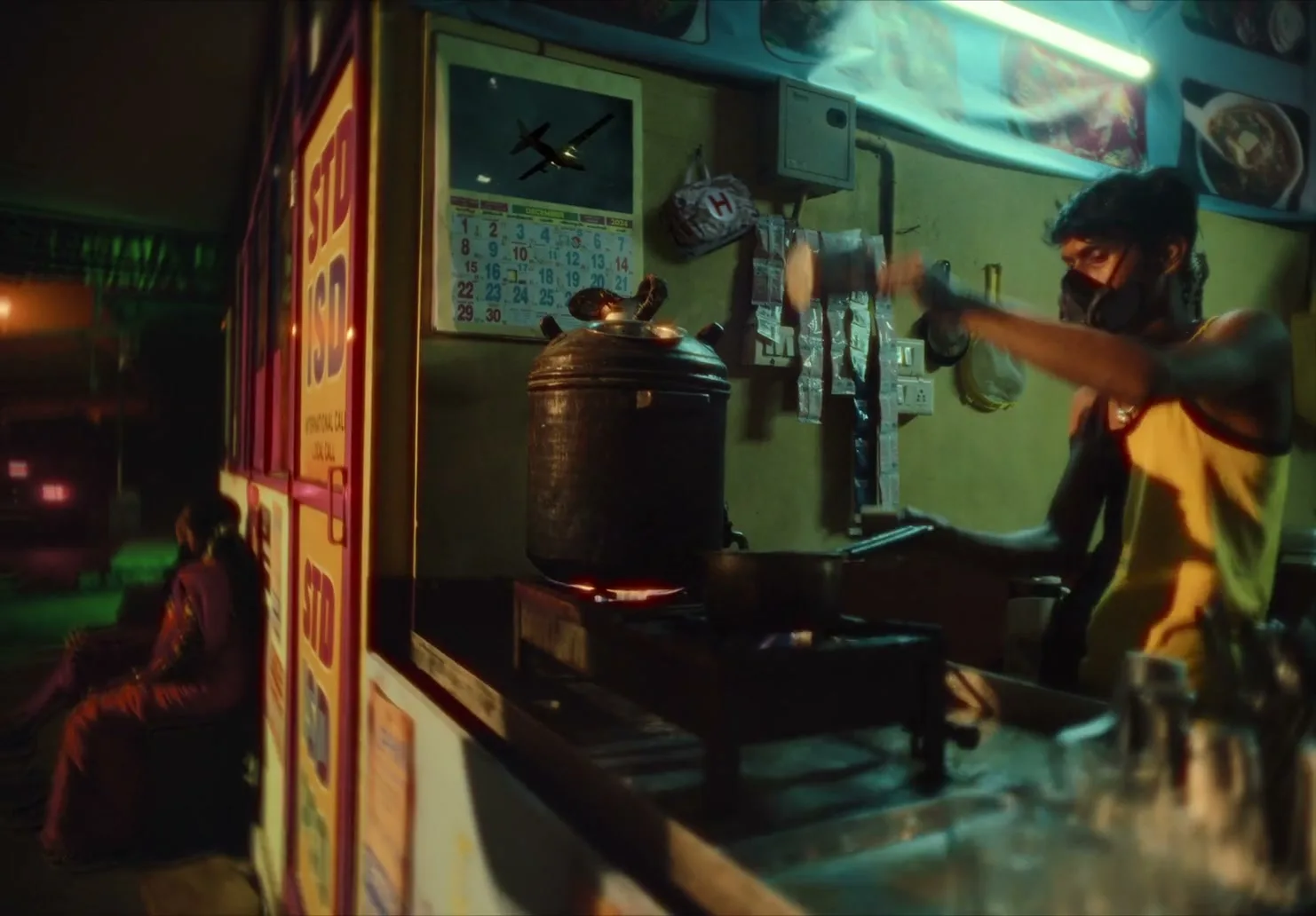 A man wearing a gas mask and yellow tank top prepares drinks at a street food stall at night, with a calendar showing an airplane on the wall.