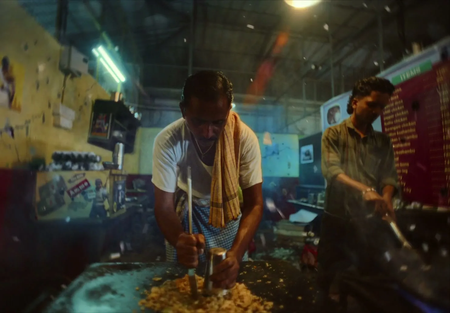 Two men cook food at a street food stall at night, with one man crushing ingredients and the other stirring a wok, as sparks fly around them.