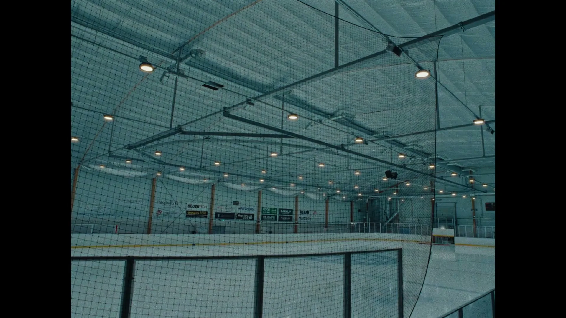 An empty ice hockey rink with protective netting and bright overhead lights is shown from a low angle, emphasizing the vastness of the arena.
