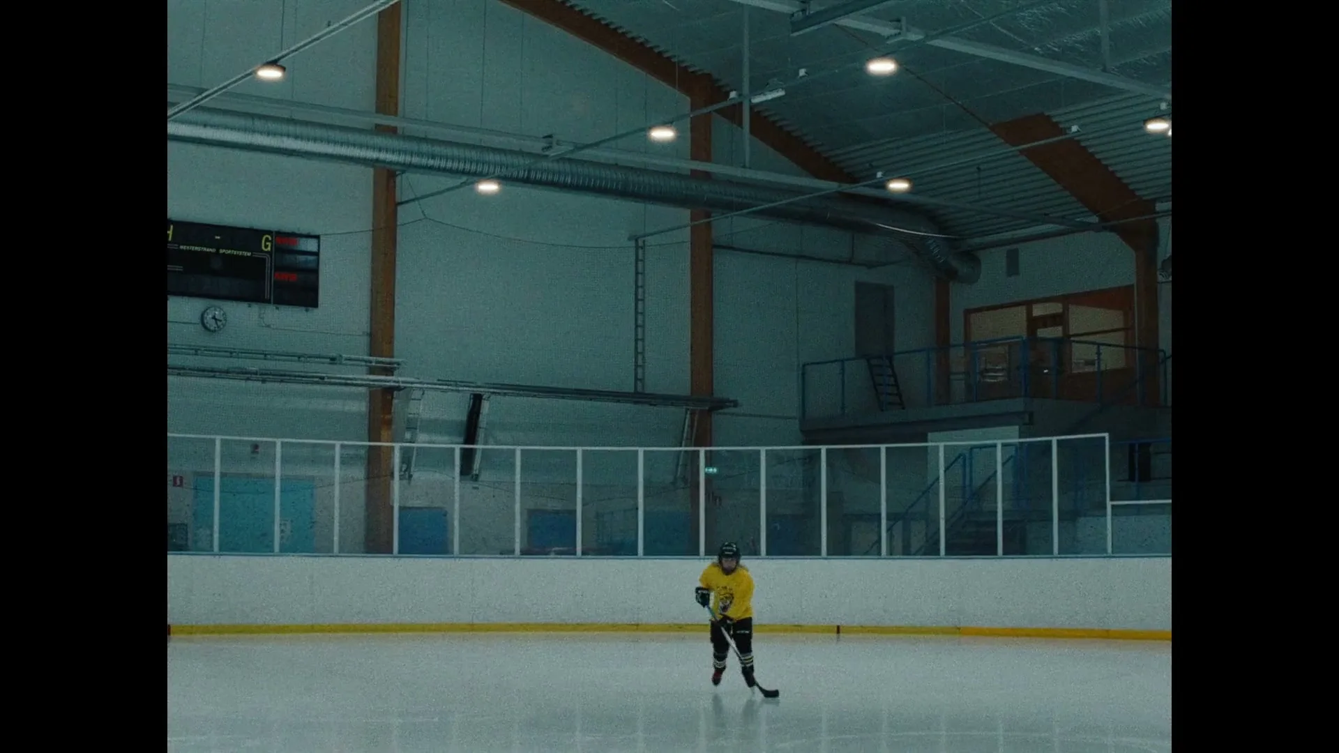 A young male ice hockey player in a yellow jersey skates across an empty ice rink in a wide shot, holding a hockey stick.