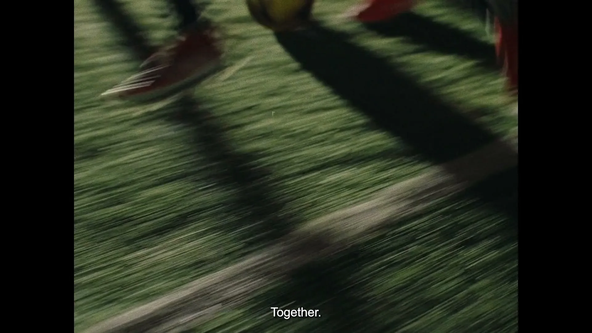 A low-angle shot captures the feet of several people running across a grassy field with a white line, suggesting a sports game.