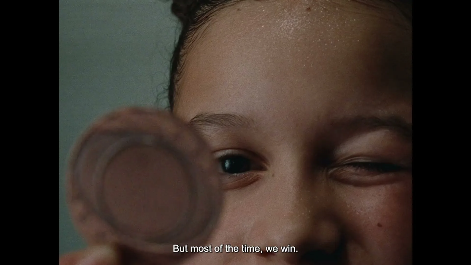 A young girl with a sweaty face winks at the camera while holding a small, round object in front of her eye.