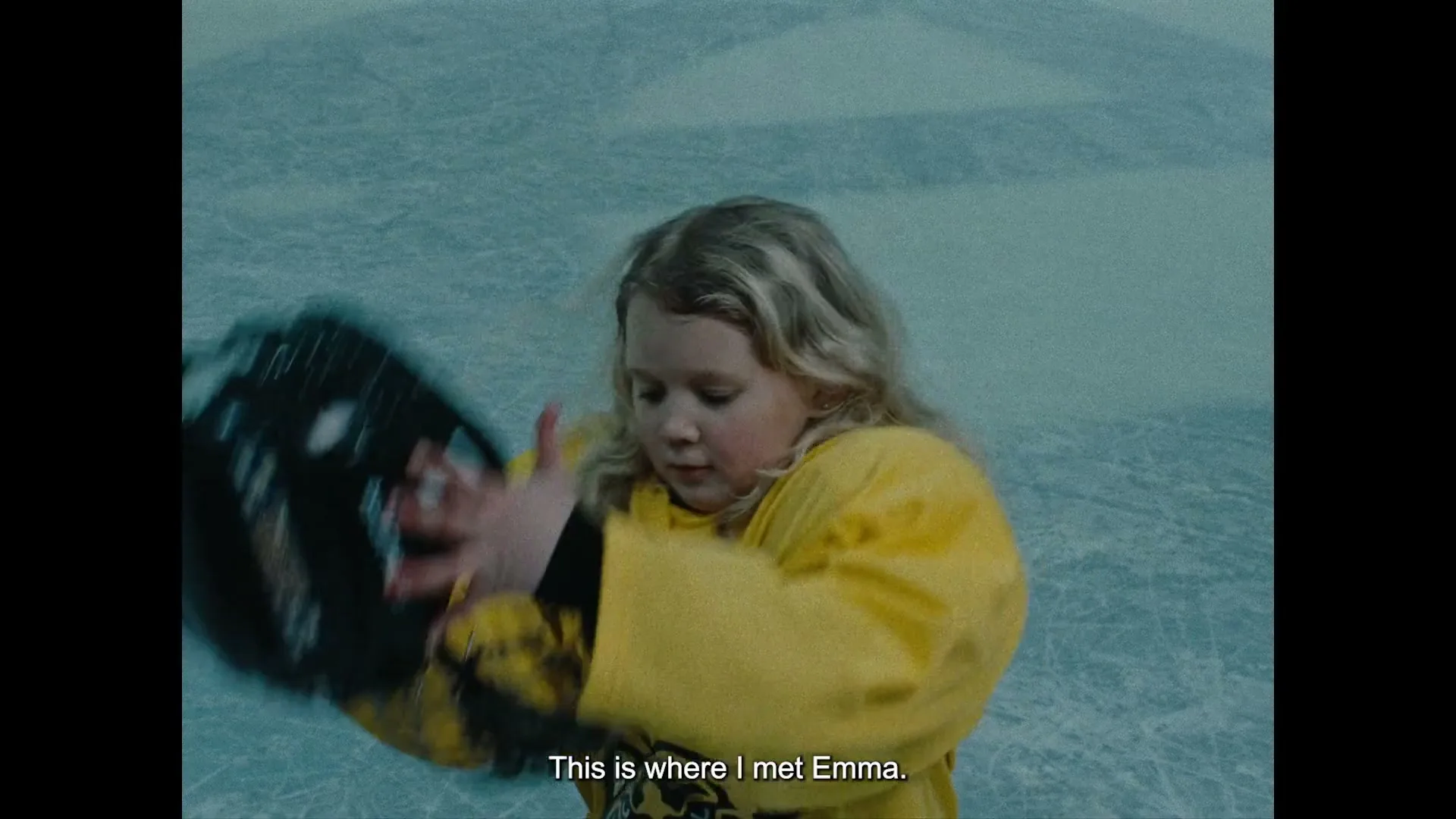 A young blonde girl in a yellow hockey jersey removes her black helmet on an ice rink.