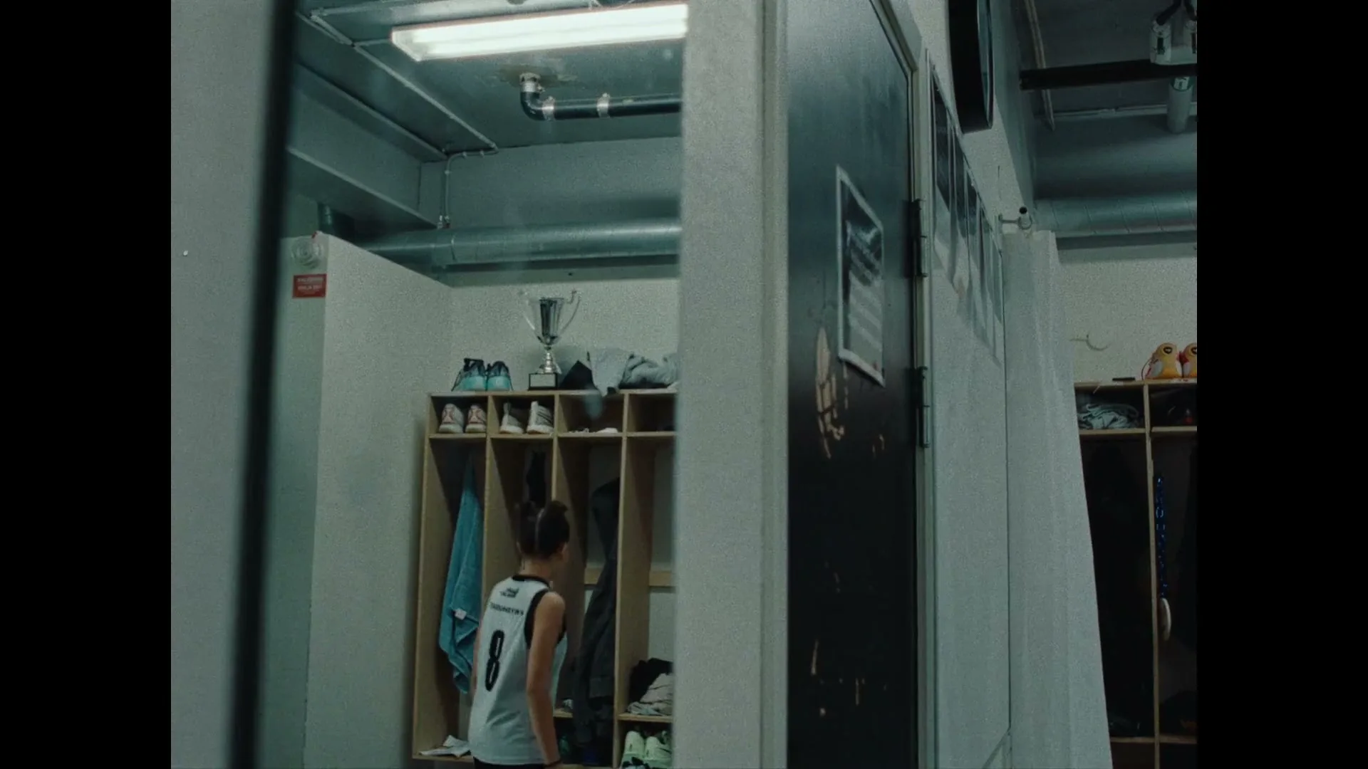 A young female basketball player walks into a locker room, sits down on a bench, and covers her face with her hands, looking distressed.