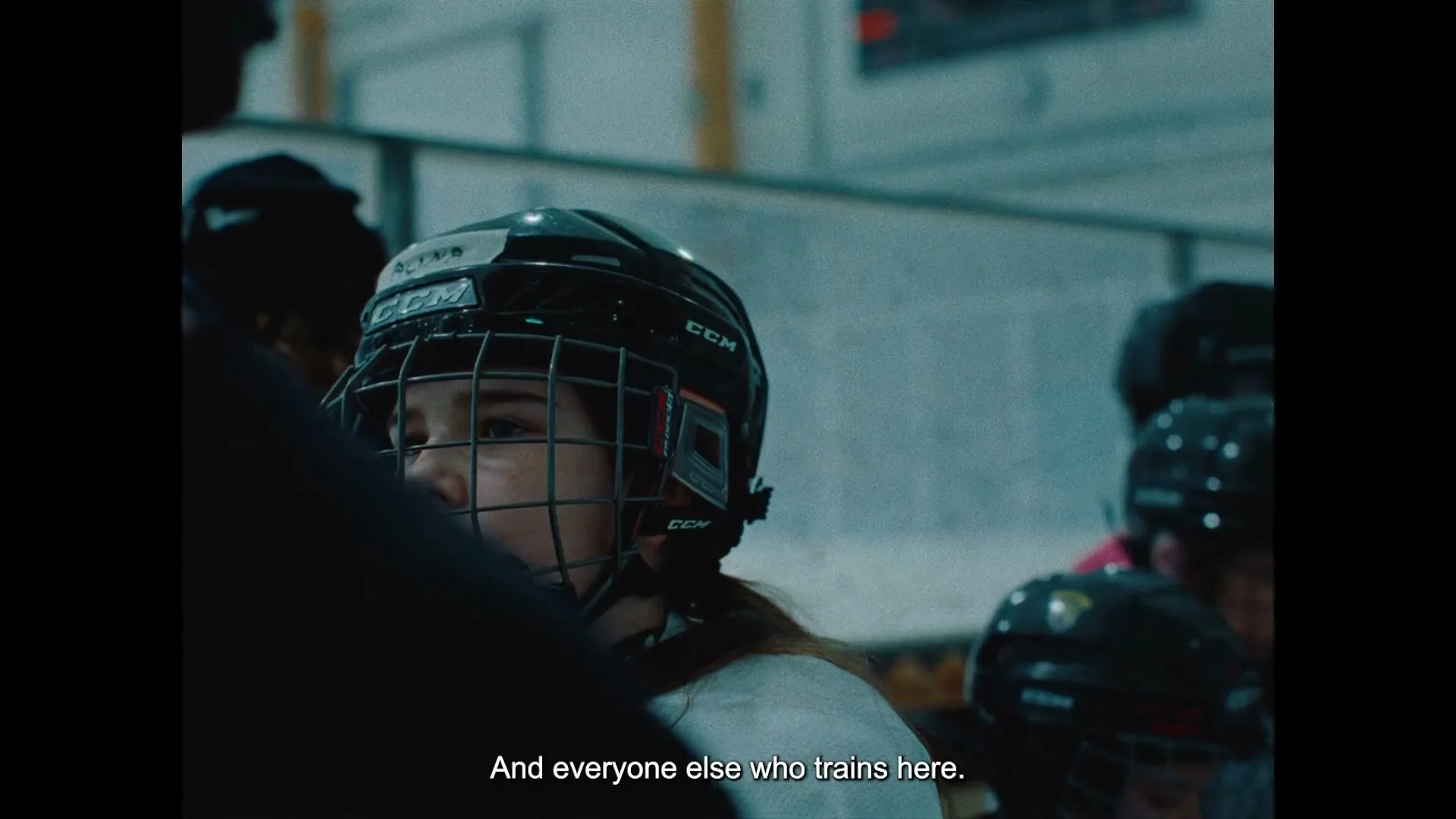 A young female ice hockey player in a black helmet and white jersey looks intently forward while sitting on a bench in an indoor ice rink.