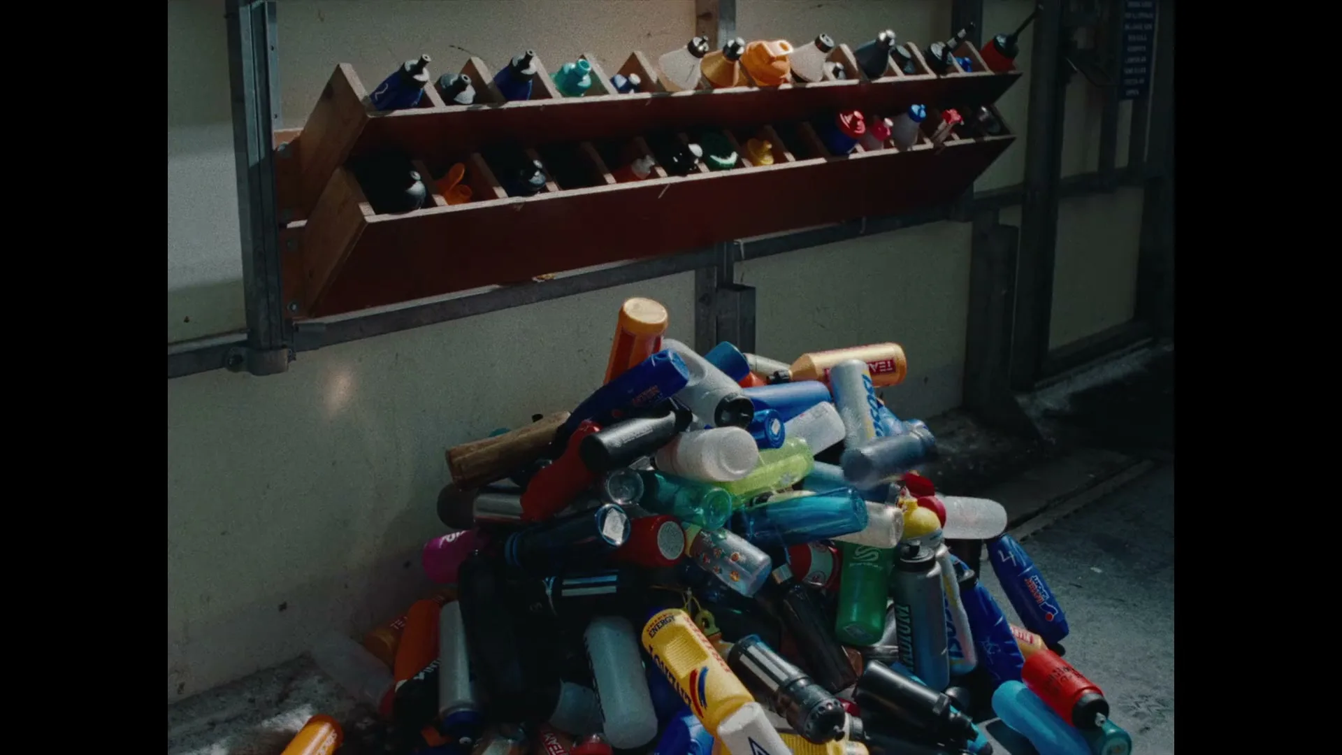 A pile of colorful plastic water bottles lies on the floor beneath a wooden rack holding more bottles in a dimly lit room.