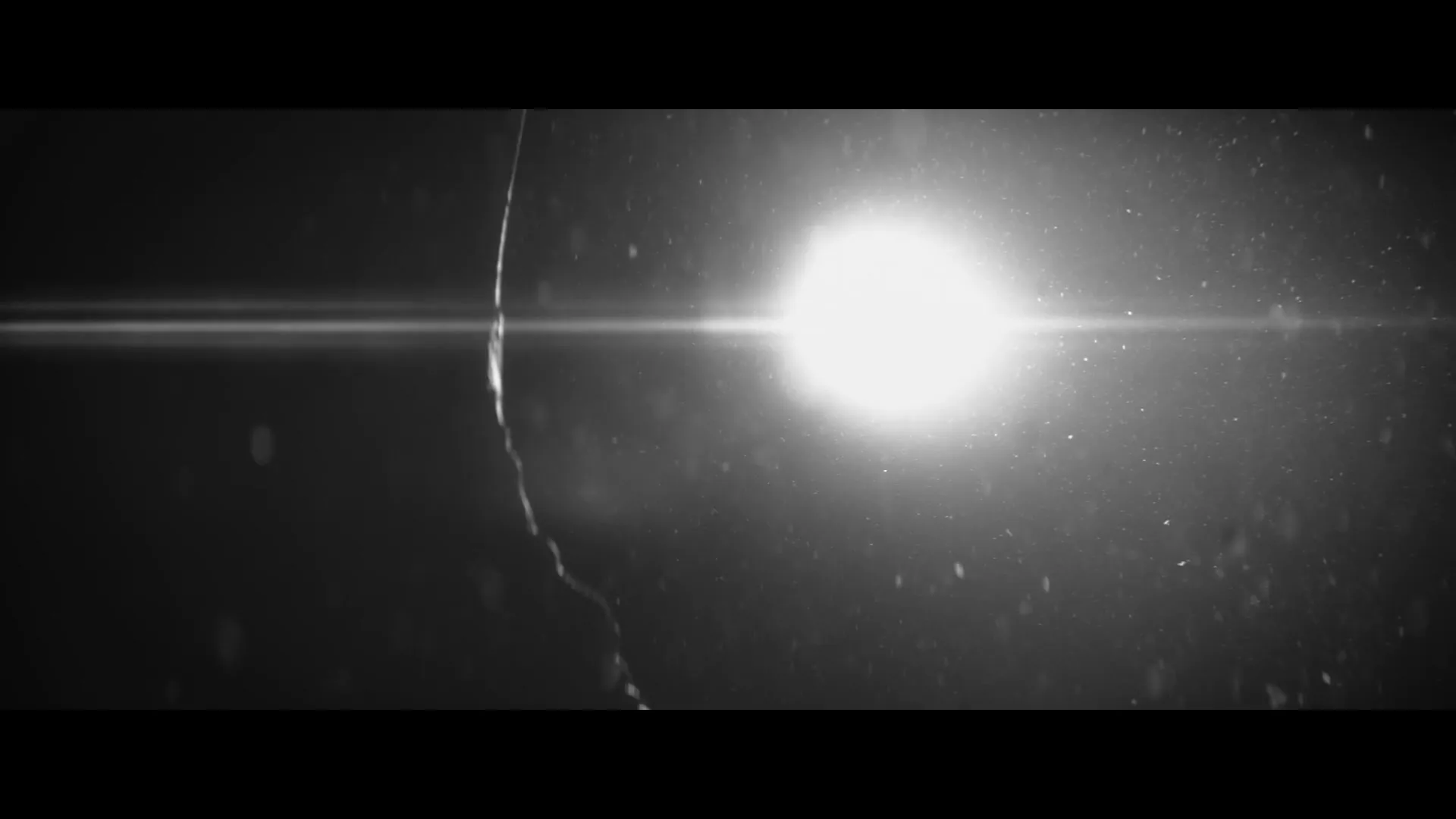 A bright light source shines through falling rain or dust particles in a dark, ambiguous space, creating a lens flare.