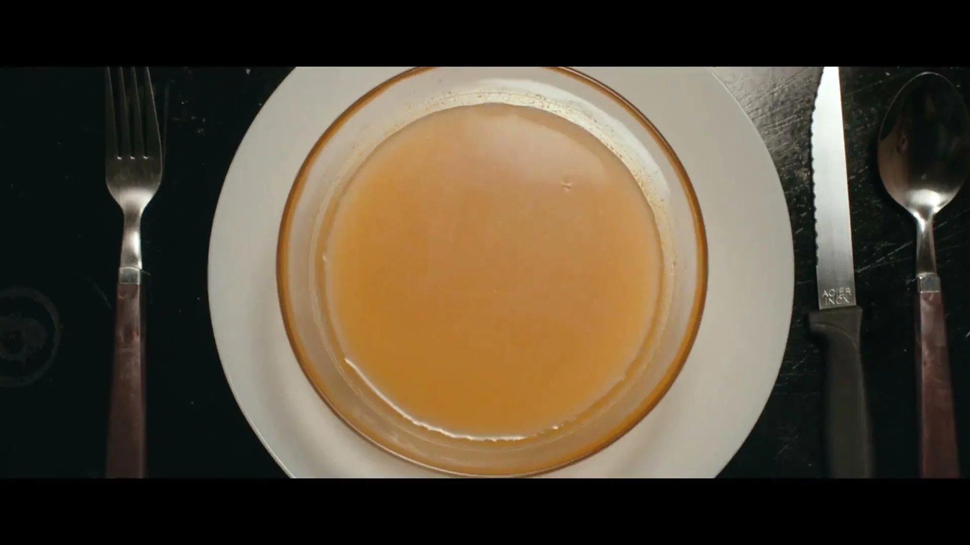 An overhead shot shows a bowl of soup on a white plate, flanked by a fork and knife, with ripples appearing in the soup.
