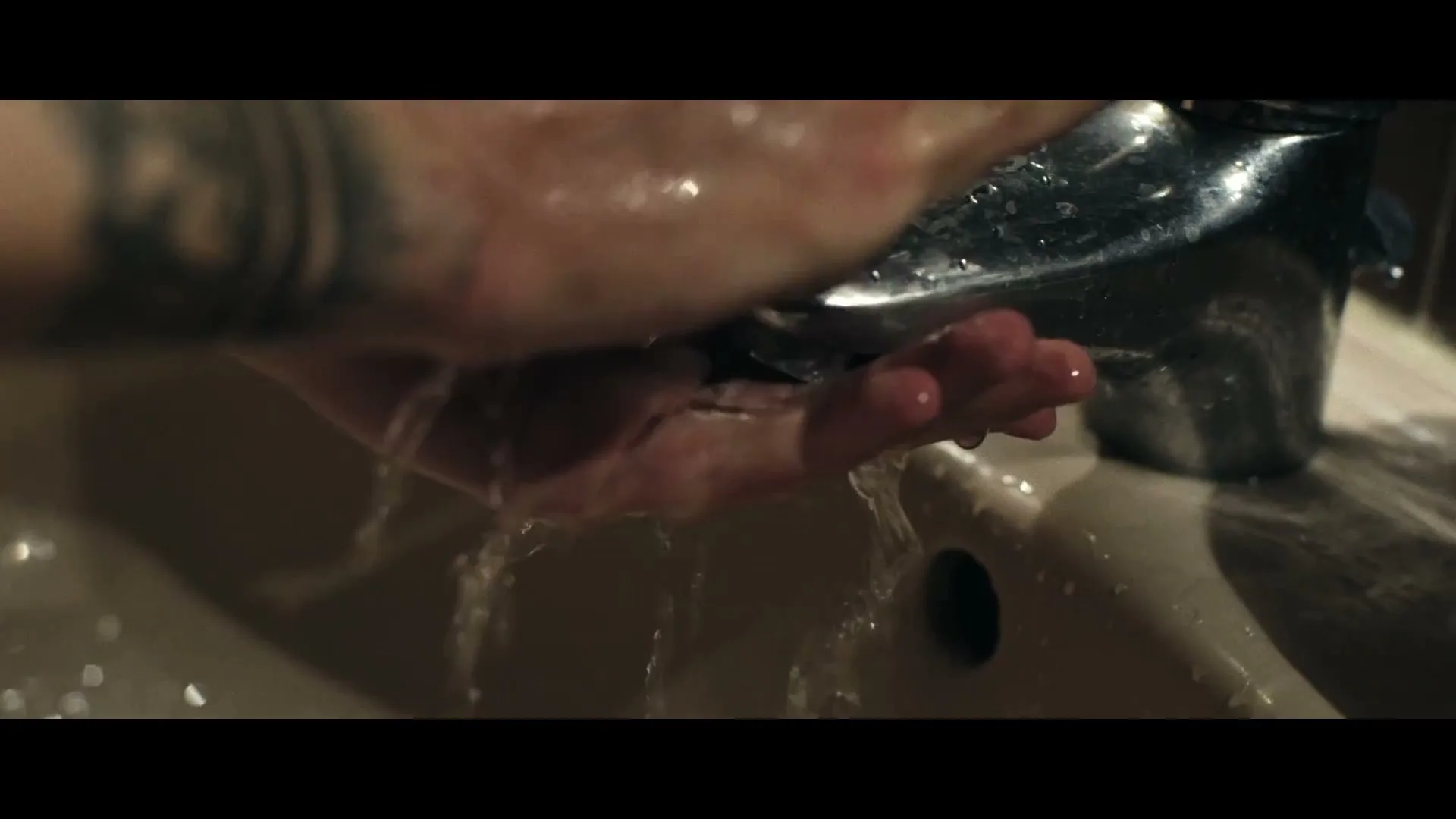 A person with tattooed arms washes their hands under a running faucet in a bathroom sink, with water splashing around.