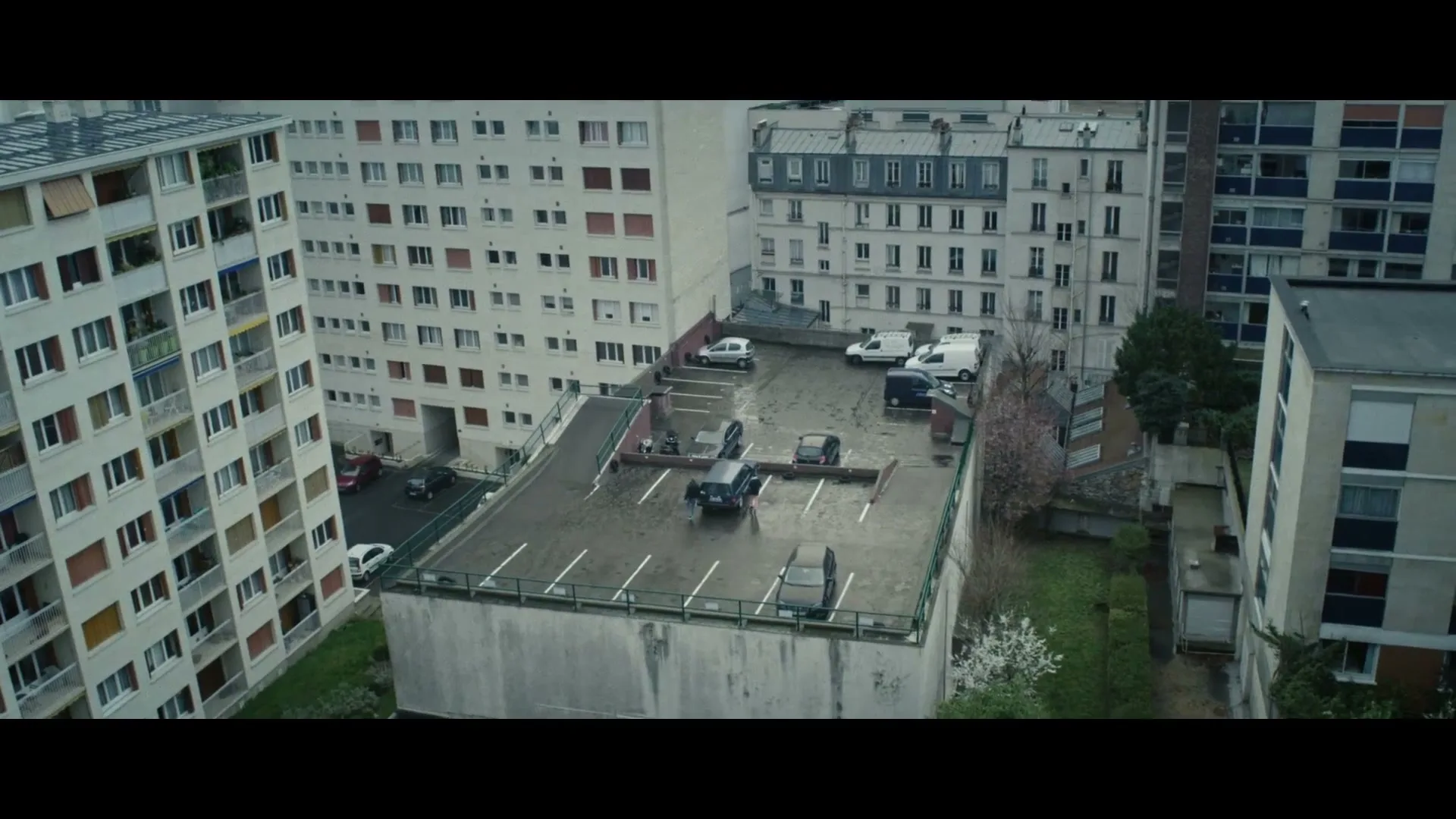 Two young women walk across a wet rooftop parking lot surrounded by apartment buildings on an overcast day.