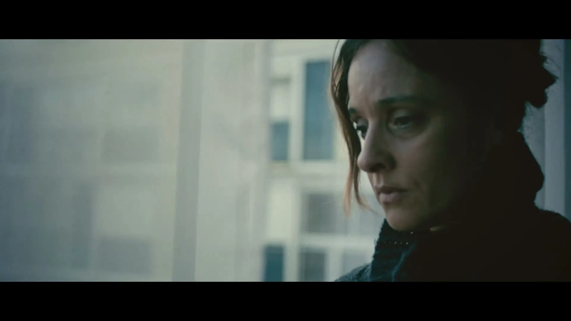 A woman with dark hair and a dark jacket looks down sadly, standing by a window in an apartment building.