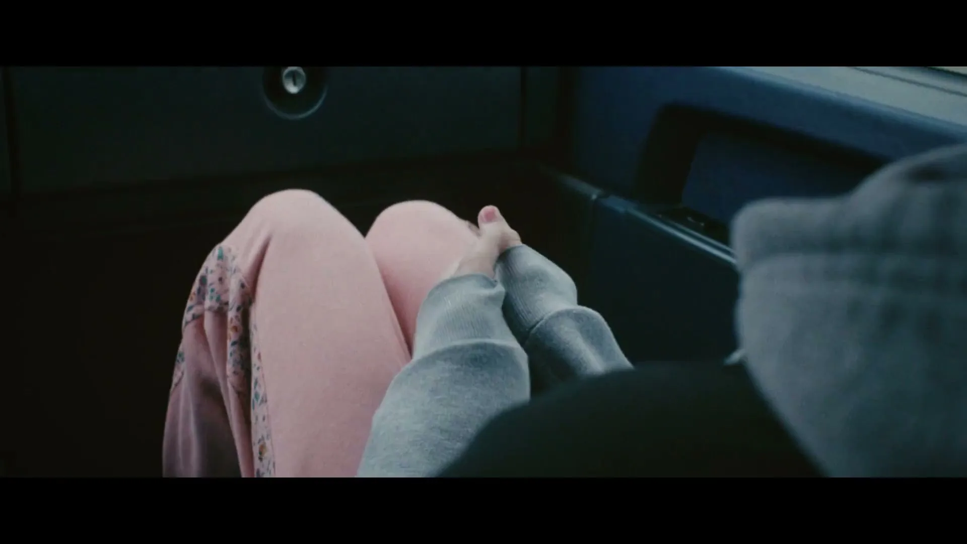 Two young women hold hands in a car, one wearing pink pants and the other a grey hoodie, as they drive during the day.