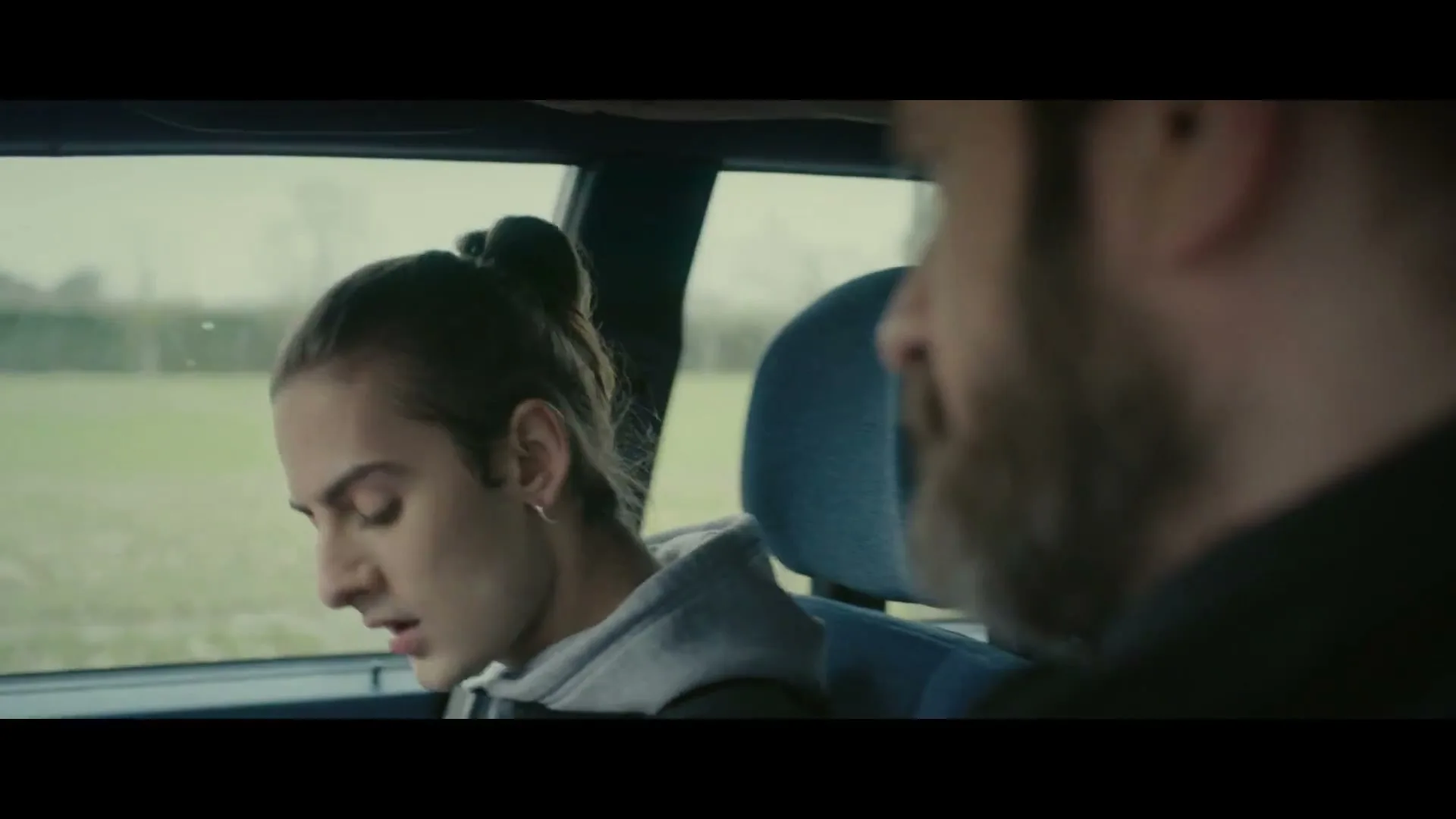 A young person with long hair tied back, wearing a grey hoodie, sits in the passenger seat of a car, looking out the window, then turns to look at the driver.