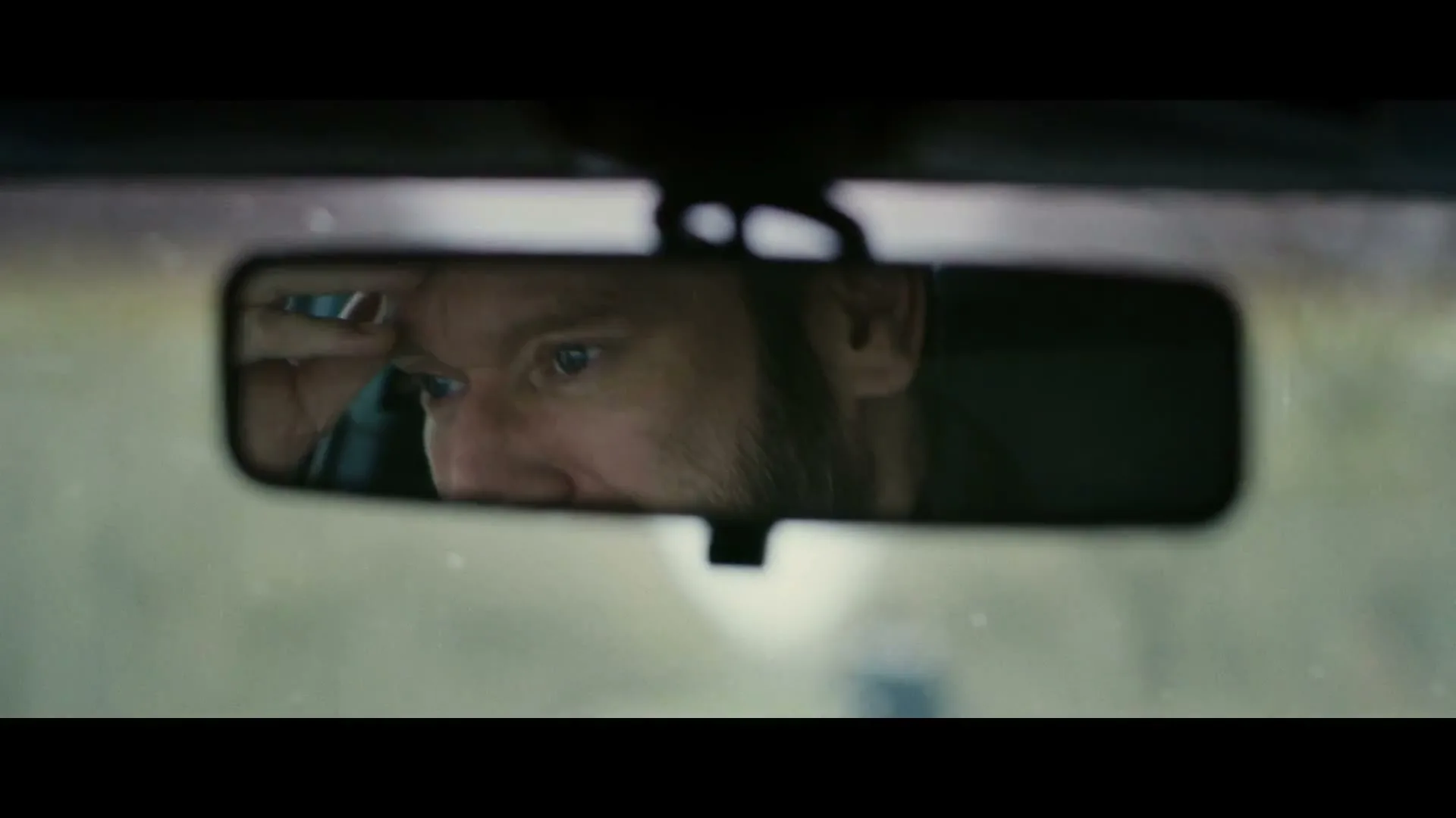 A man with a beard is seen in a car's rearview mirror, looking distressed and rubbing his forehead while driving on a road.
