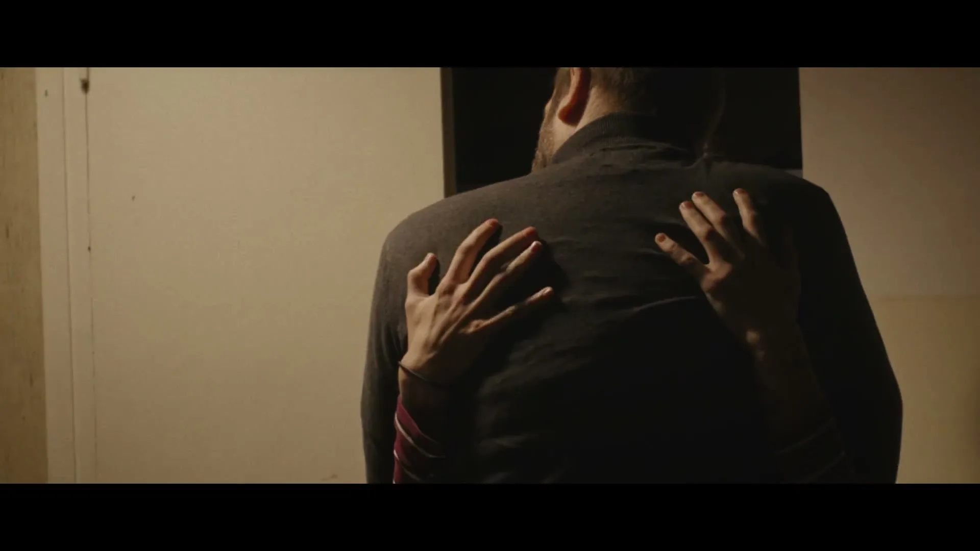 A person with painted fingernails embraces another person from behind, their hands resting on the other's back in a dimly lit room.