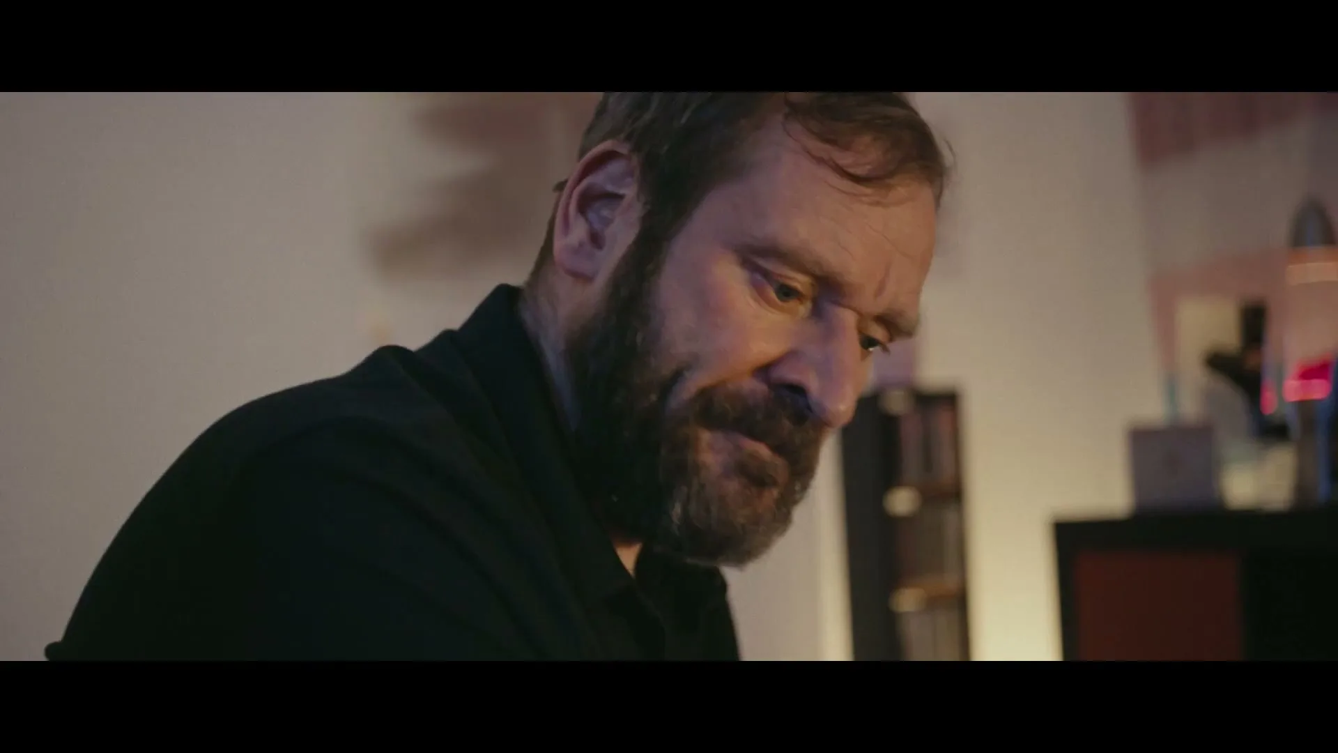 A bearded man in his 40s, wearing a black shirt, looks down with a sad expression in a dimly lit room.
