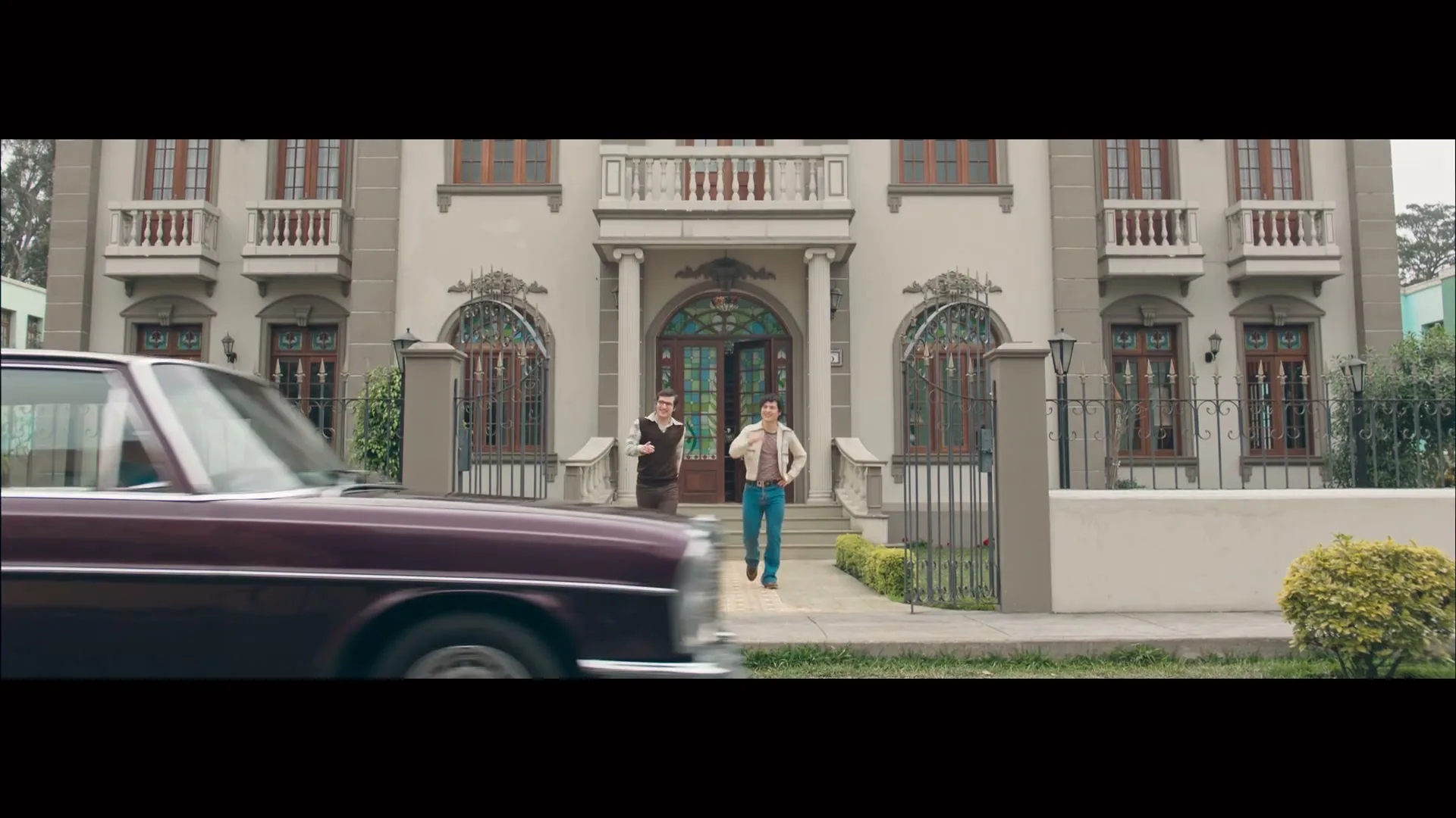 Two men in 70s attire walk out of a grand house as a vintage car drives past them on a suburban street.