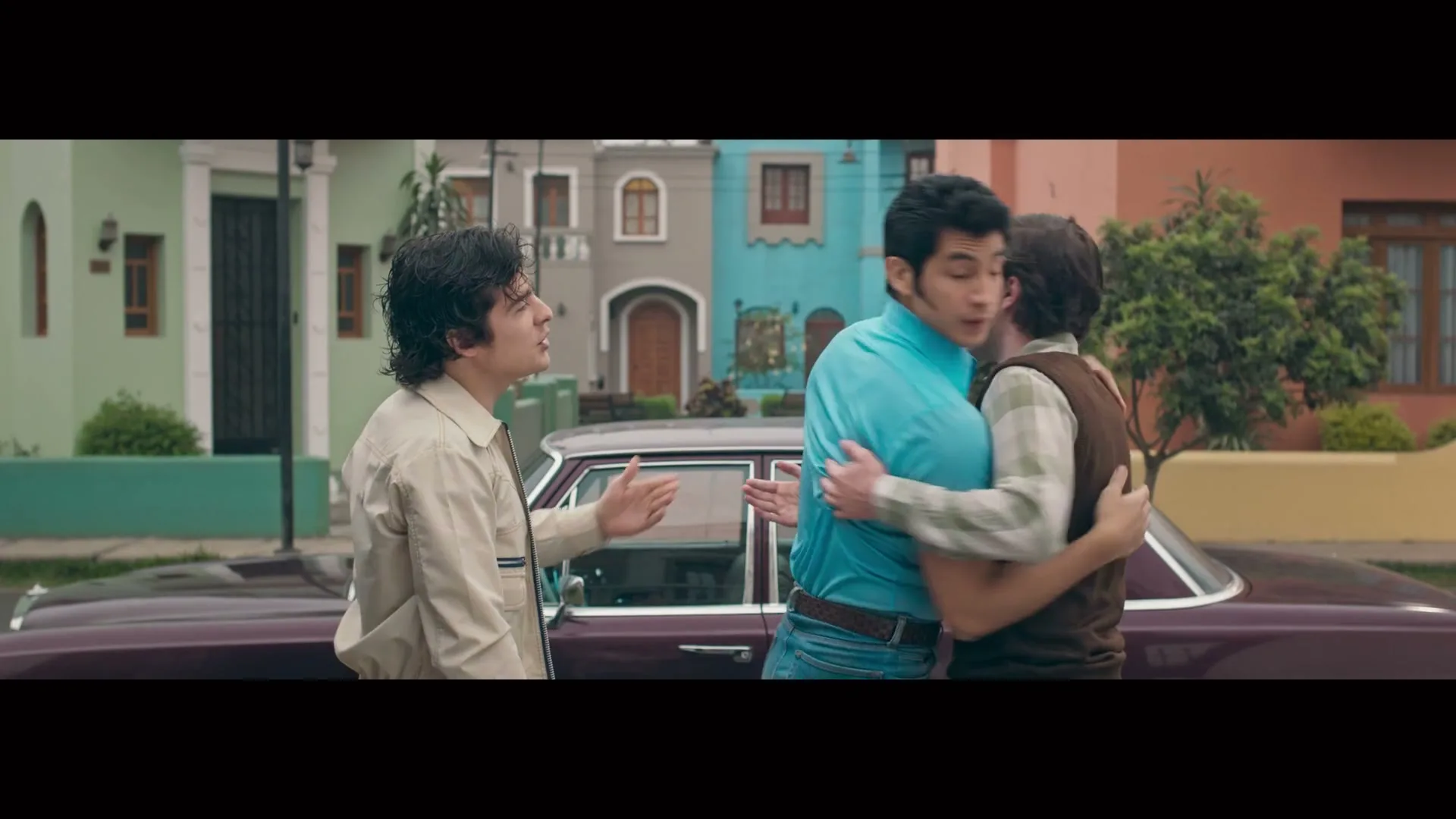 Three men stand on a street next to a vintage car, talking and gesturing, with colorful houses in the background.