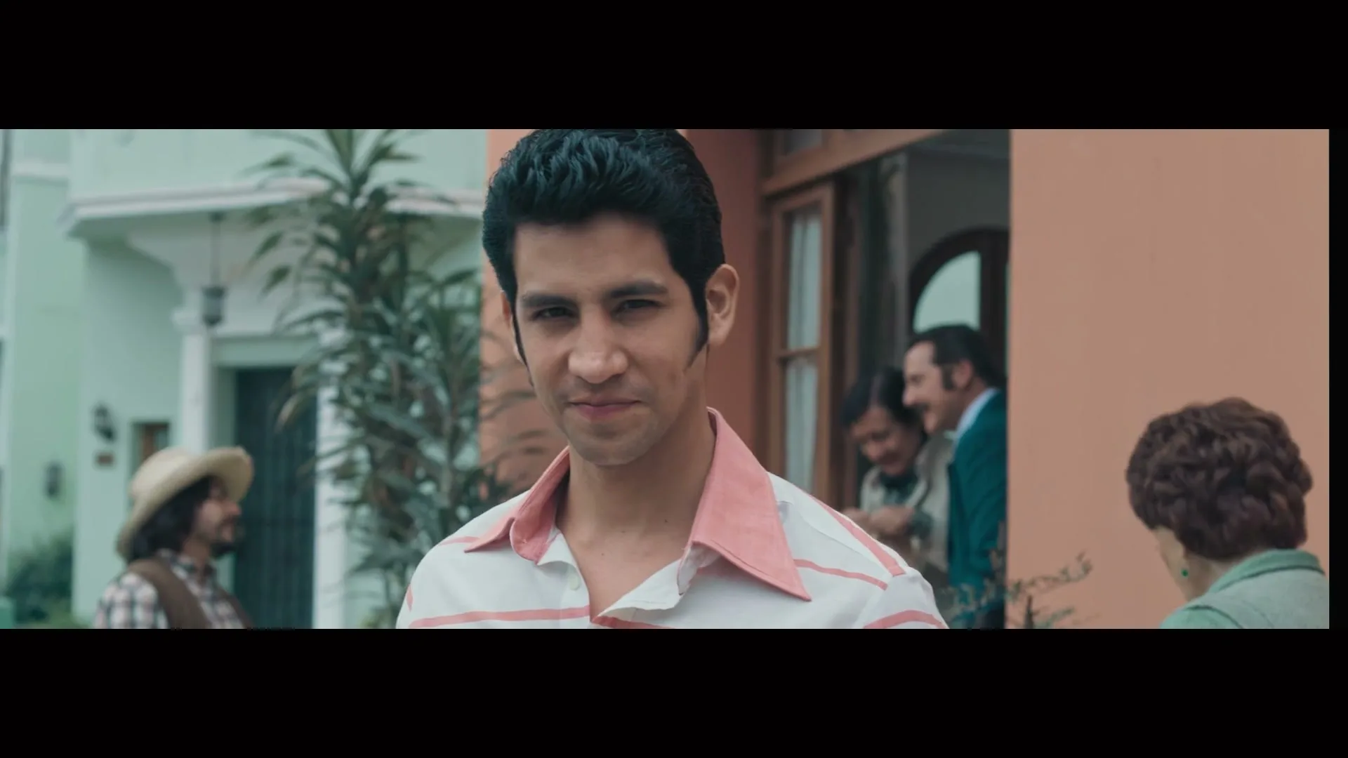 A young man with dark hair and a pink-collared shirt smiles at the camera, while other people are blurred in the background.