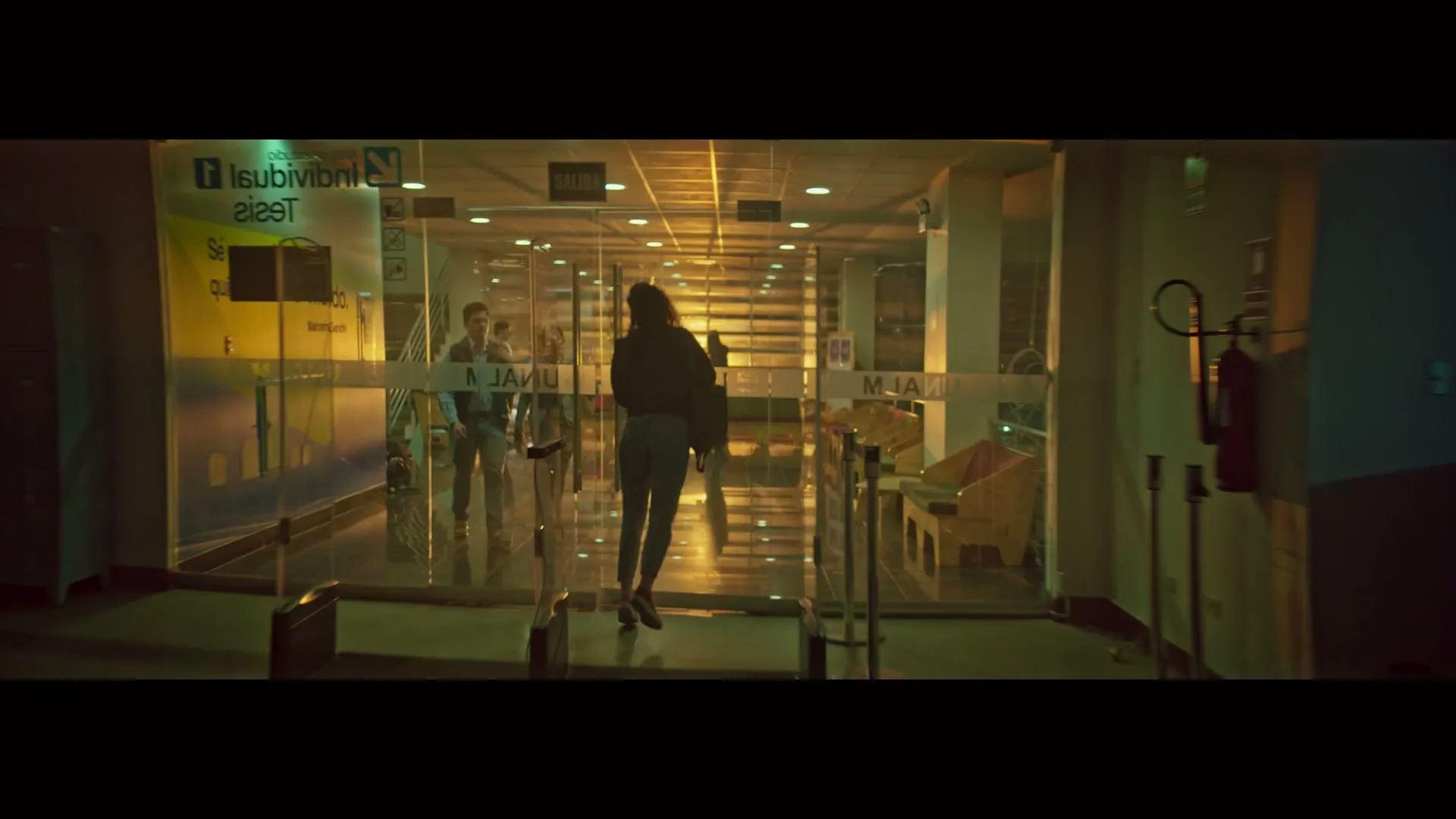 A young woman with dark hair, wearing a dark jacket and jeans, runs towards a glass door in a dimly lit building, carrying a bag.