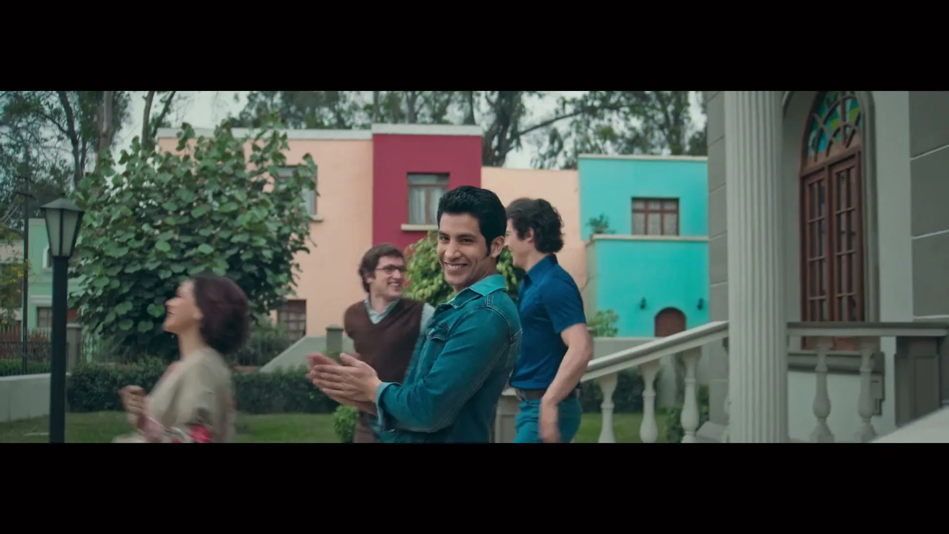 A group of friends, including a man in a denim jacket, walk down a path in a colorful neighborhood, smiling and clapping.