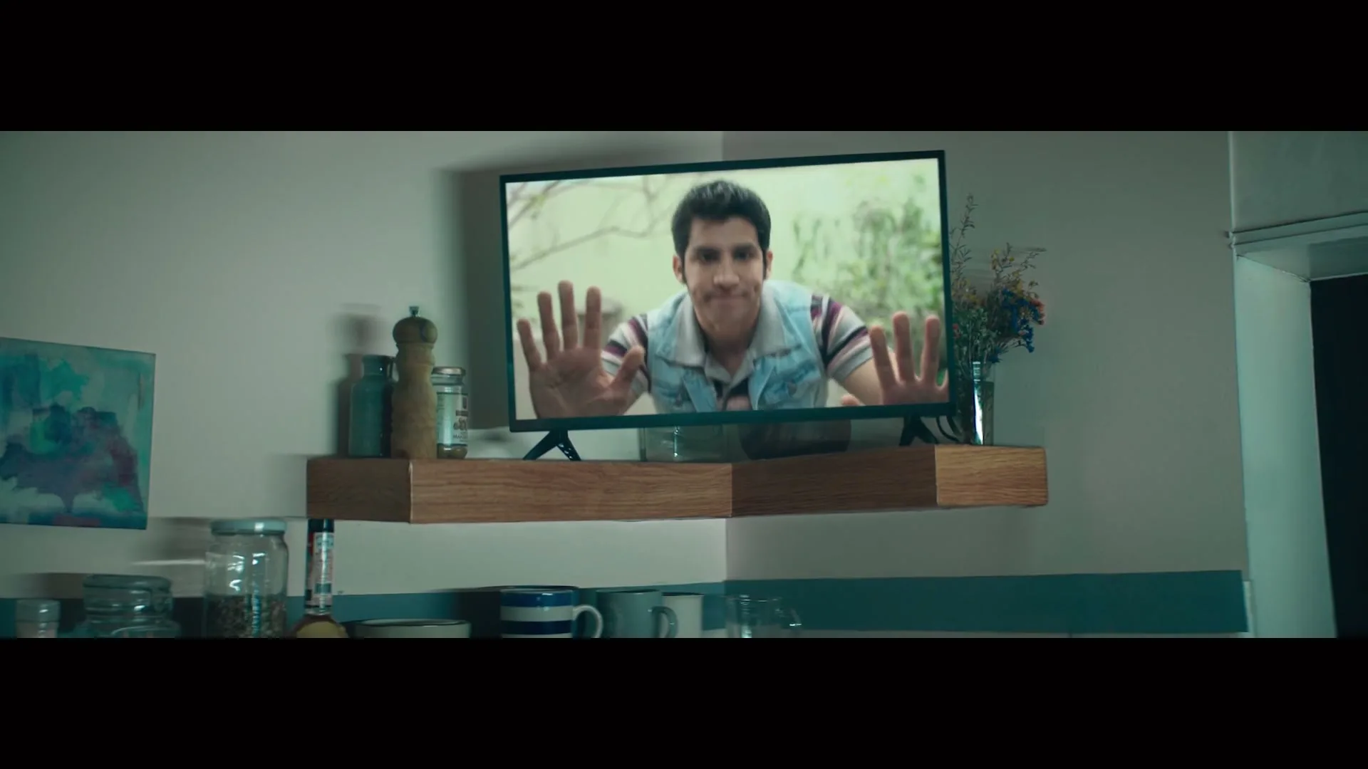 A young man with dark hair and a striped shirt is seen on a TV screen, pushing his hands against the screen as if trying to escape, in a kitchen.
