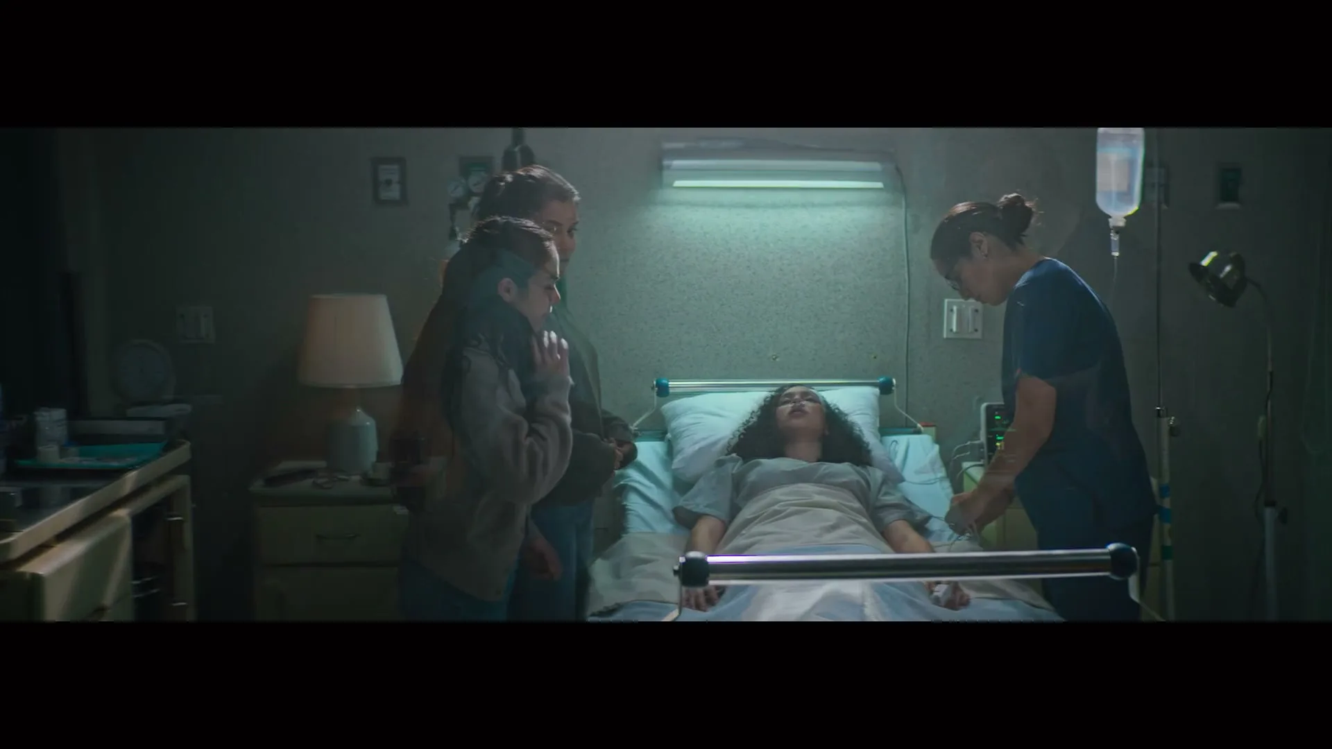 A young woman lies in a hospital bed, surrounded by family and a nurse, in a dimly lit hospital room.