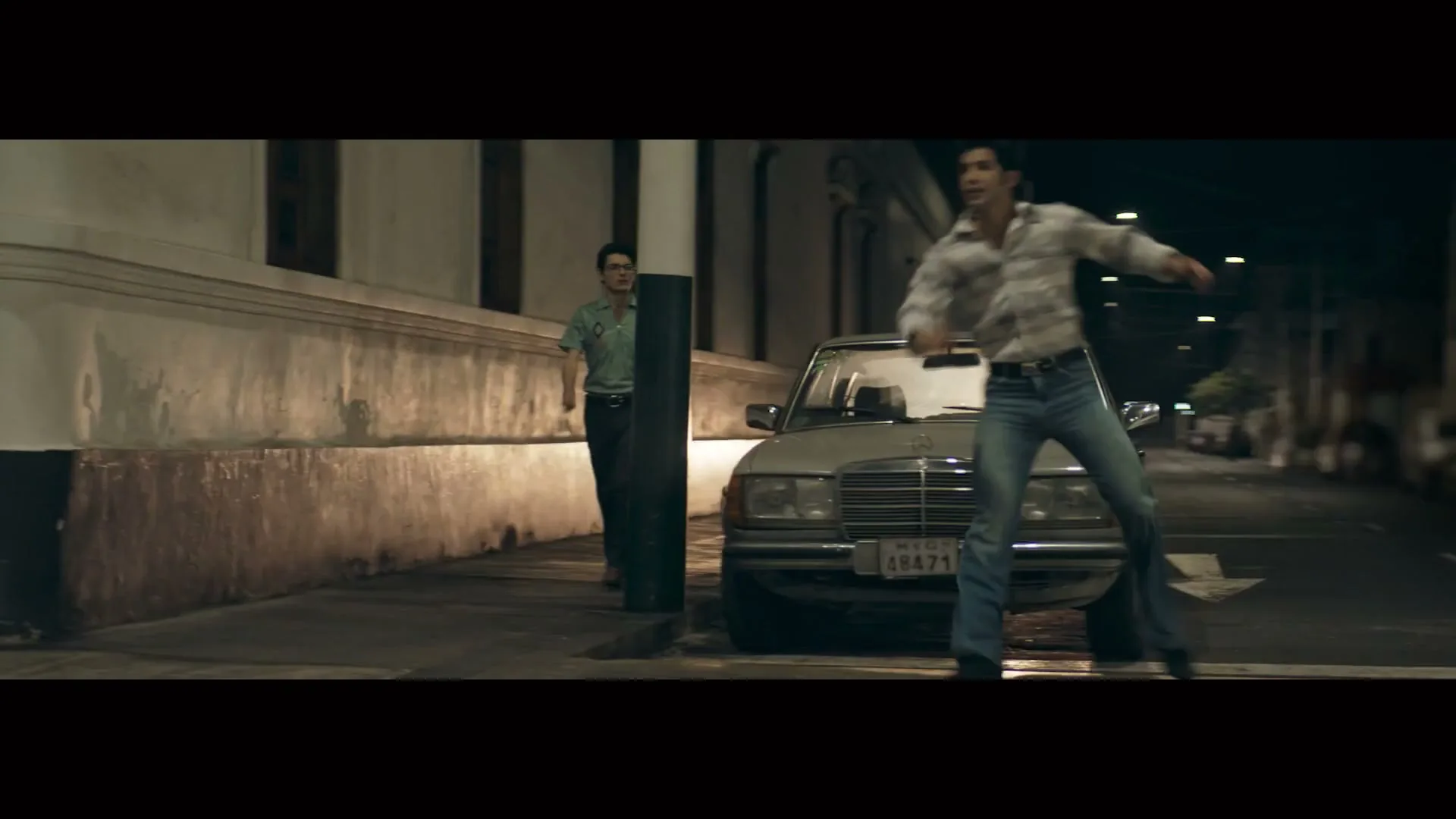 Two men in retro clothing are on a dimly lit street at night, one running and the other standing by a parked car.