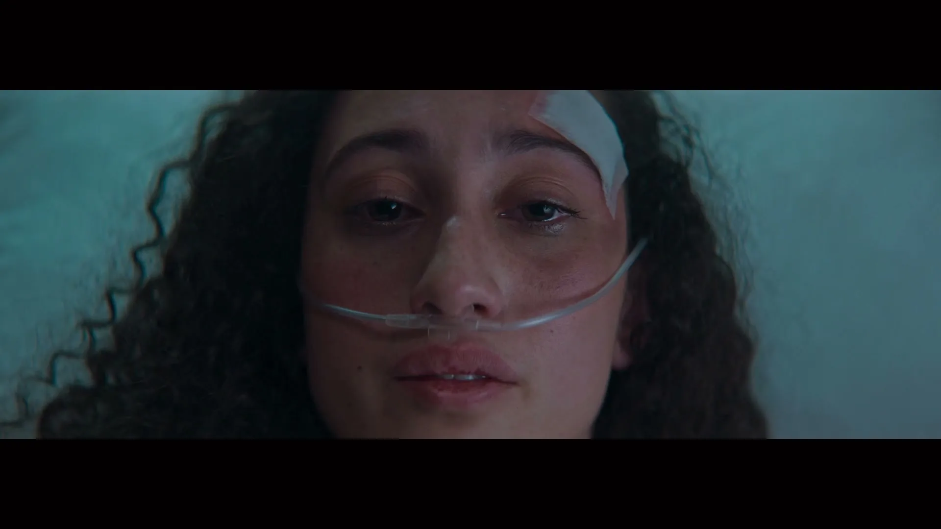 A young woman with curly hair and a bandage on her forehead lies in a hospital bed, with a nasal cannula, looking up with tears in her eyes.
