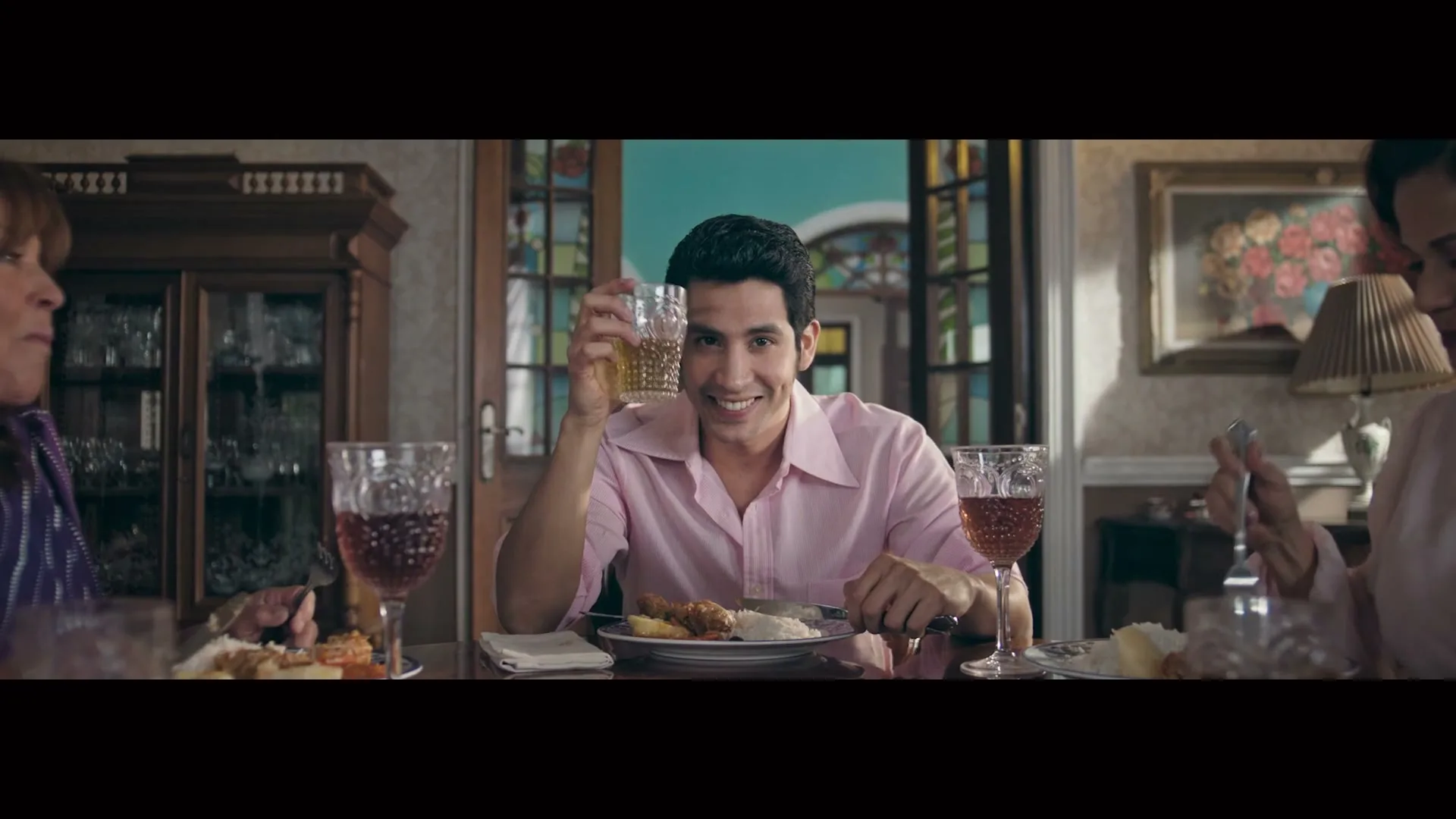 A man in his 20s, wearing a pink shirt, smiles at the camera while holding a drink at a dinner table with family.
