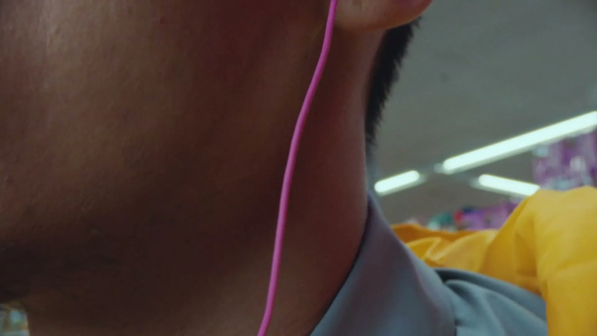 A man with a mustache and pink earbuds talks on the phone while walking through a brightly lit grocery store aisle.