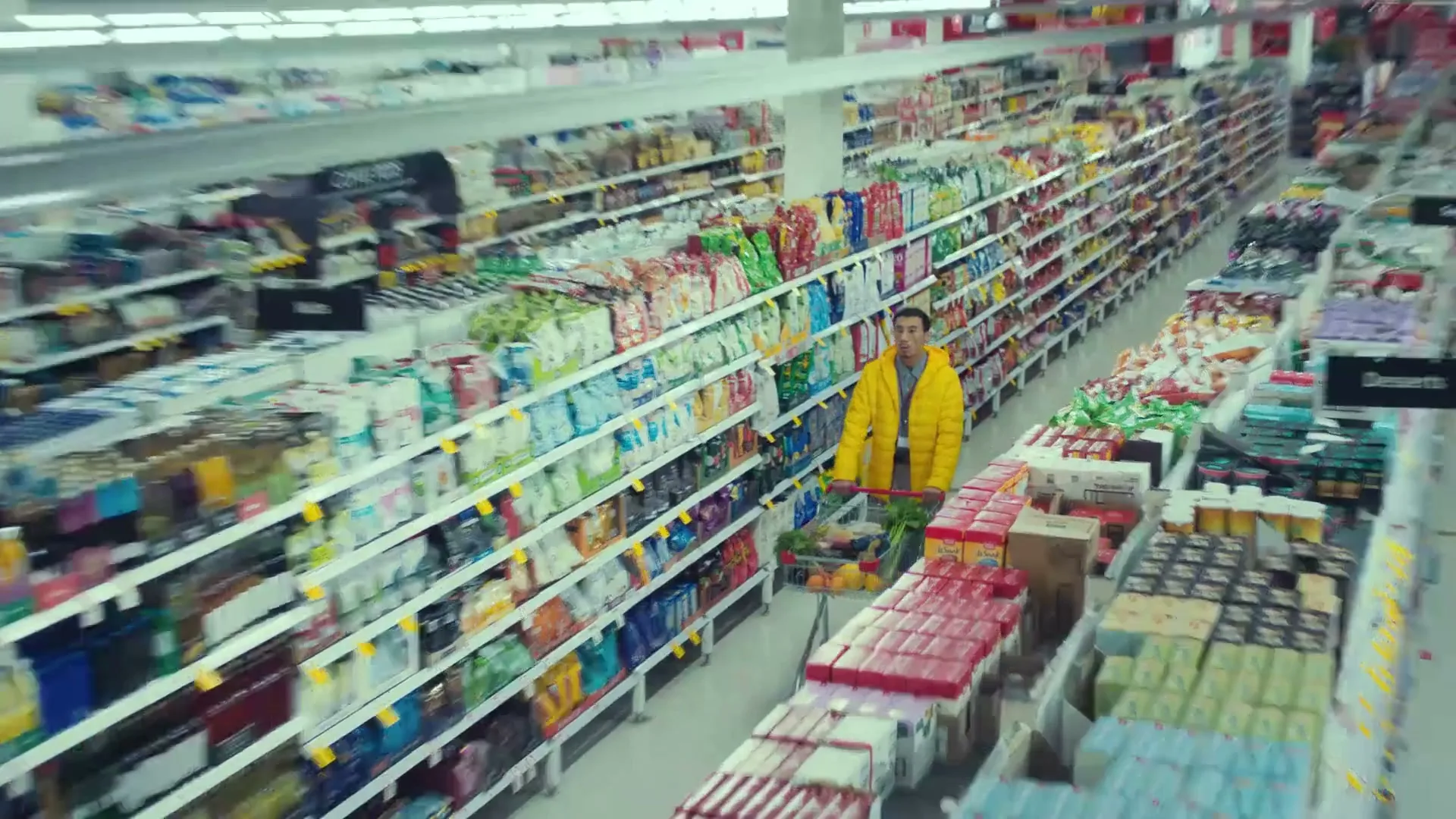 A man in a bright yellow puffer jacket pushes a shopping cart filled with groceries down a long aisle in a brightly lit supermarket.