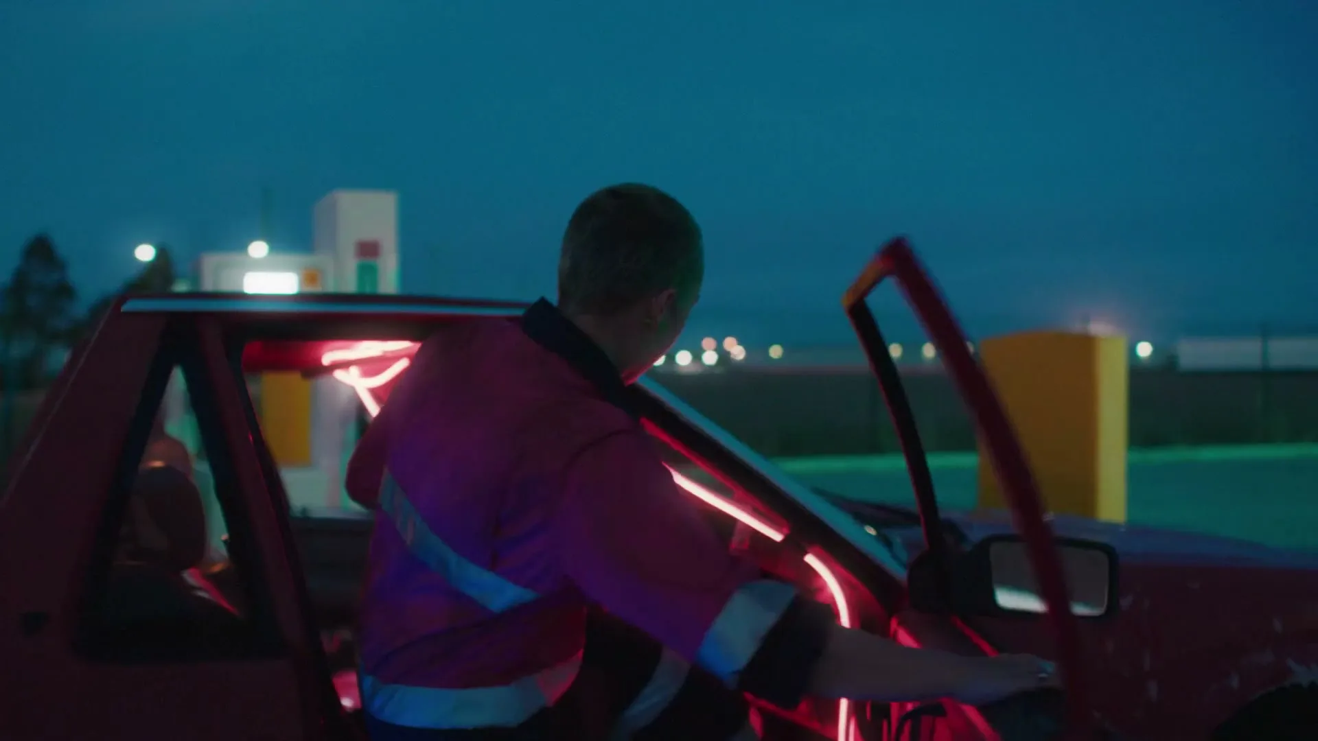 A young person with a buzzcut, wearing a pink work shirt, walks towards a red car with neon lights, then gets in and looks at the camera at a gas station at dusk.
