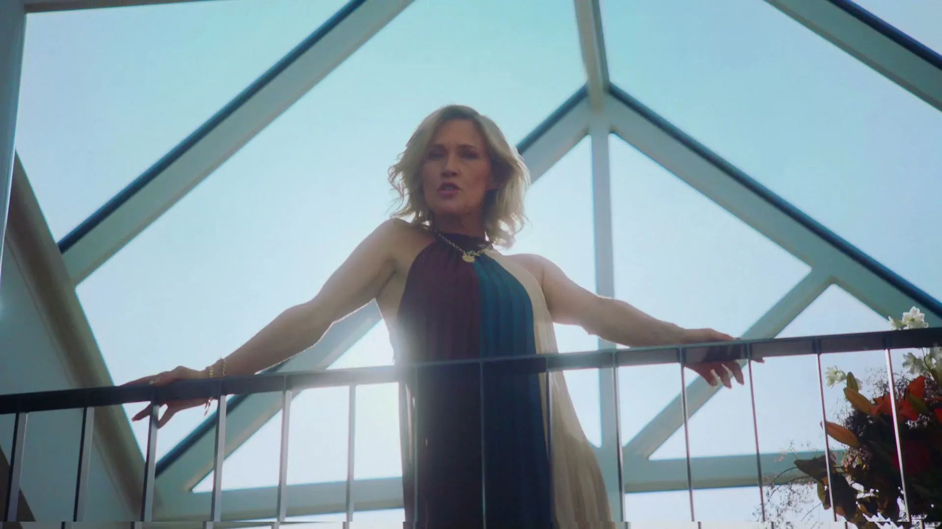 A blonde woman in a colorful dress leans on a balcony railing, looking down with a confident expression in a modern, sunlit interior.